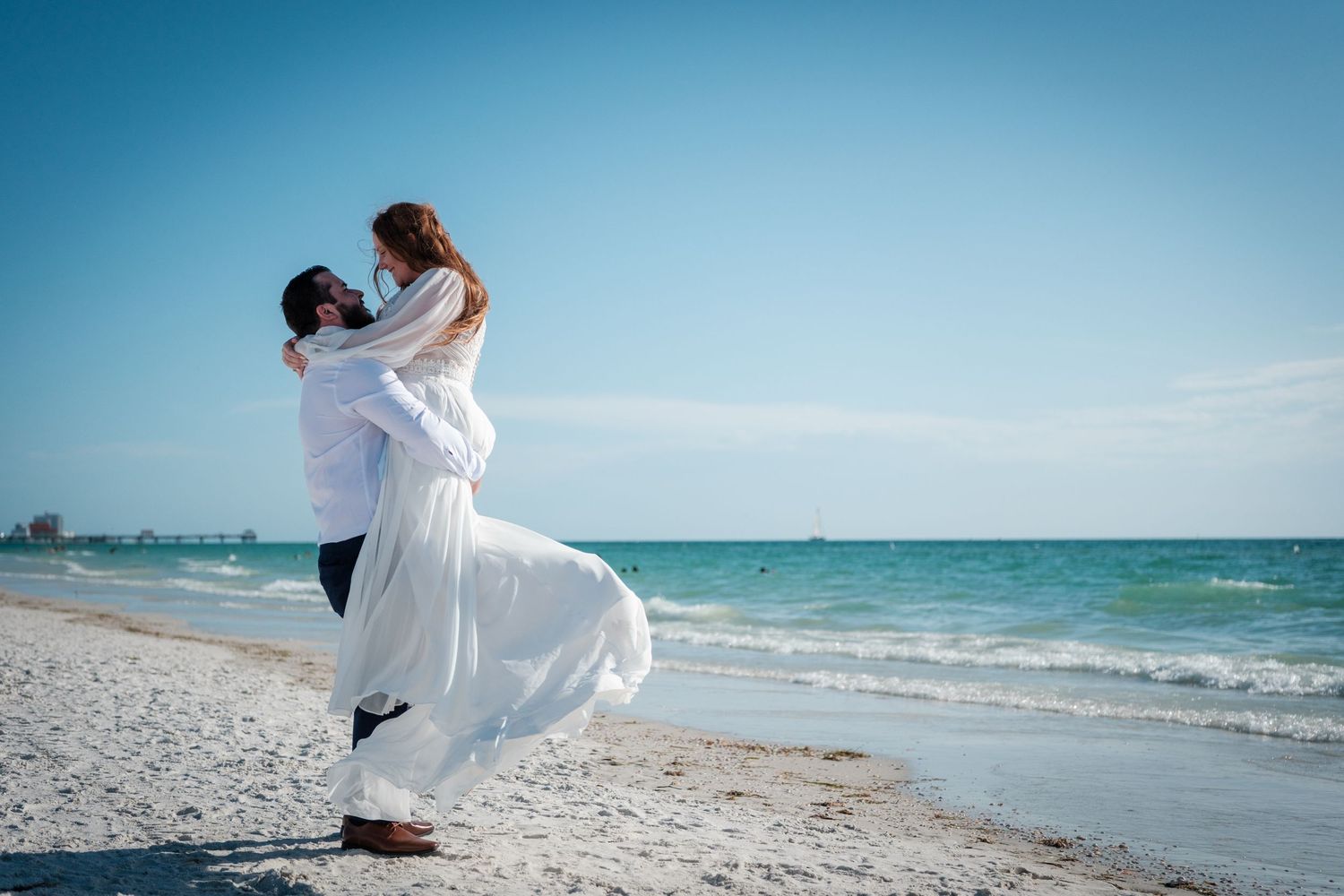 Couple in white attire sharing a romantic embrace on a sunny beach with turquoise ocean waves in the background.