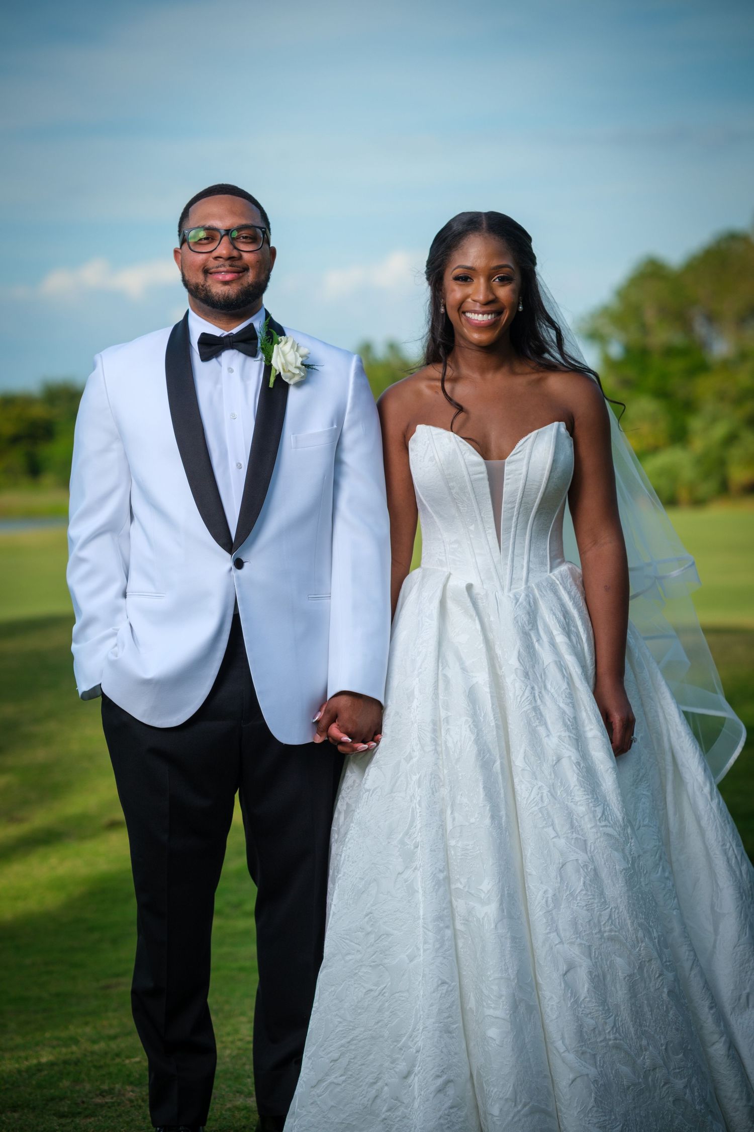 Smiling newlywed couple in white wedding attire holding hands outdoors on a green lawn under a blue sky.