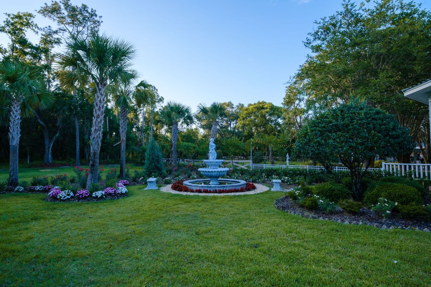 An elegant tiered fountain centerpiece in a manicured garden with palm trees and purple flowers at twilight.