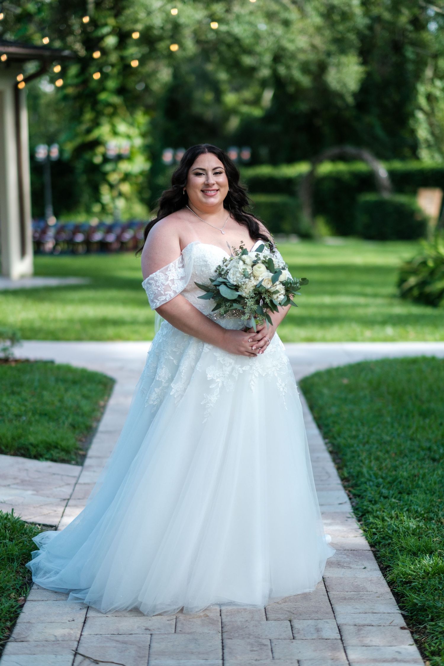 Bride in off-shoulder white ballgown walks along stone pathway surrounded by green landscaping.