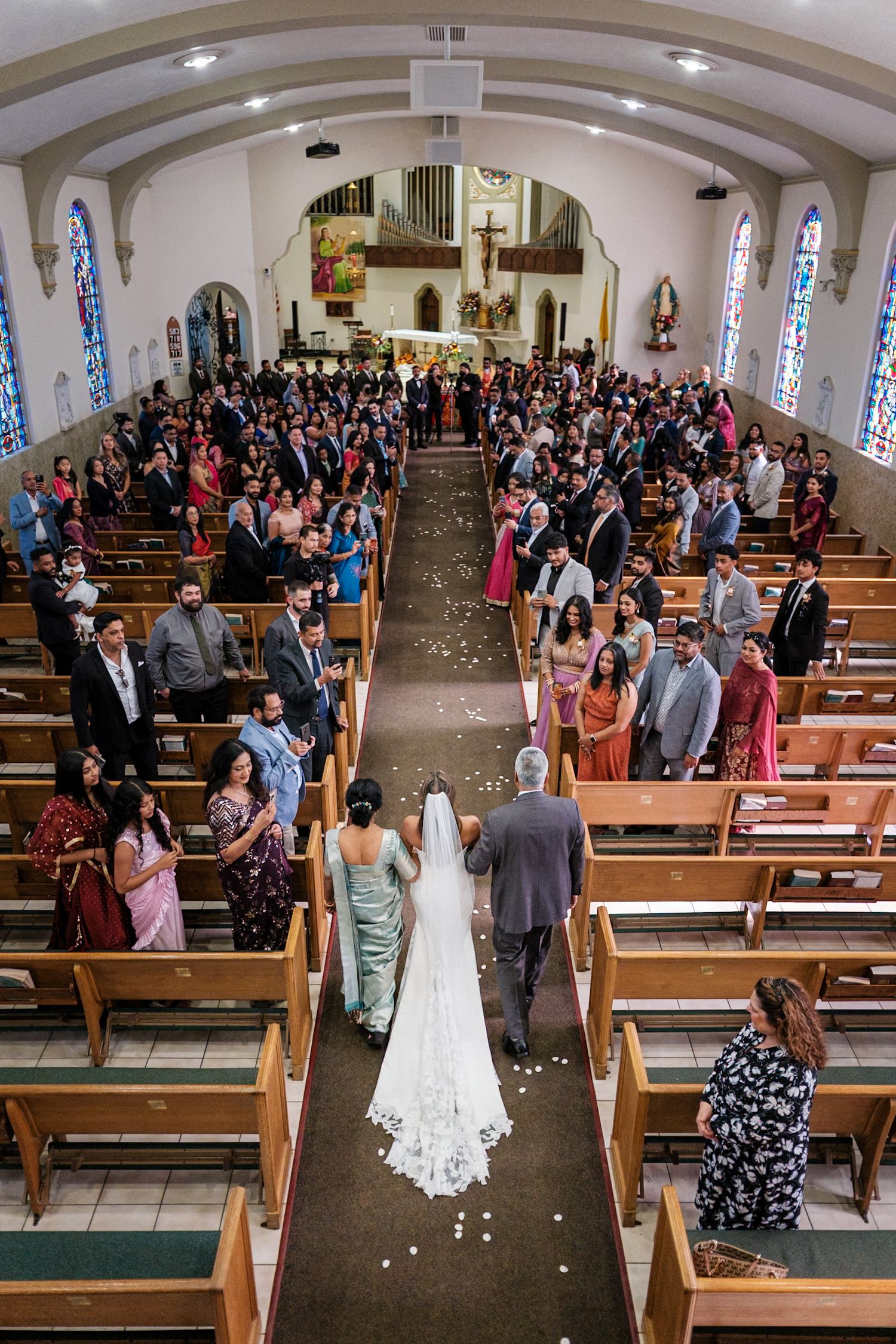 Aerial view of a traditional church wedding ceremony with guests standing in wooden pews along a flower-petaled aisle.