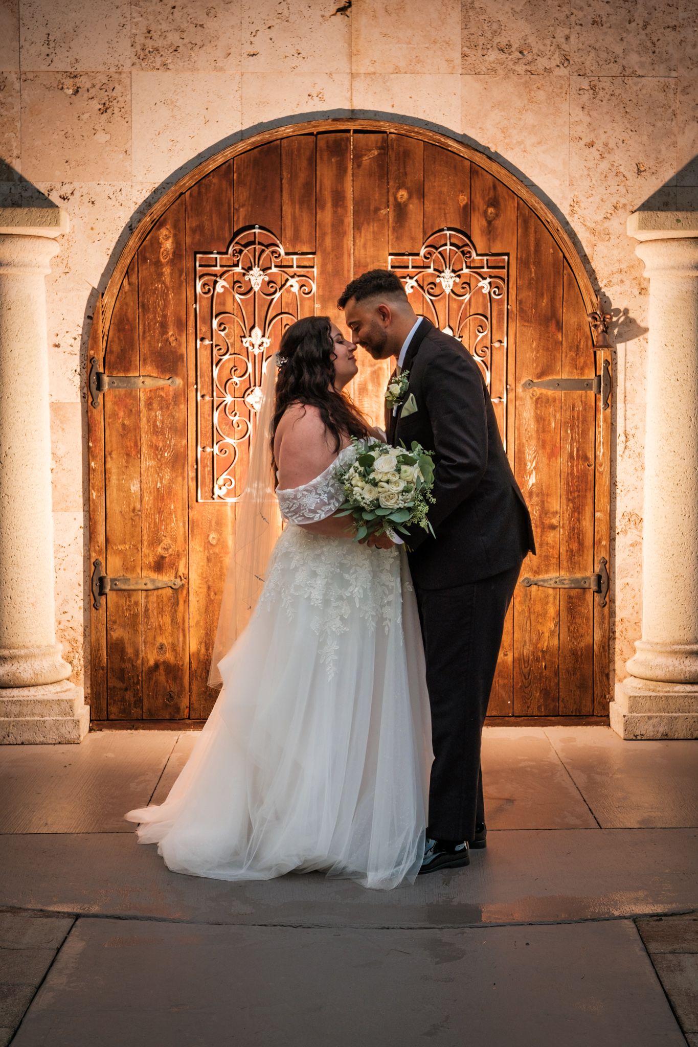 A romantic wedding scene against rustic wooden doors and stone columns in warm sepia lighting.