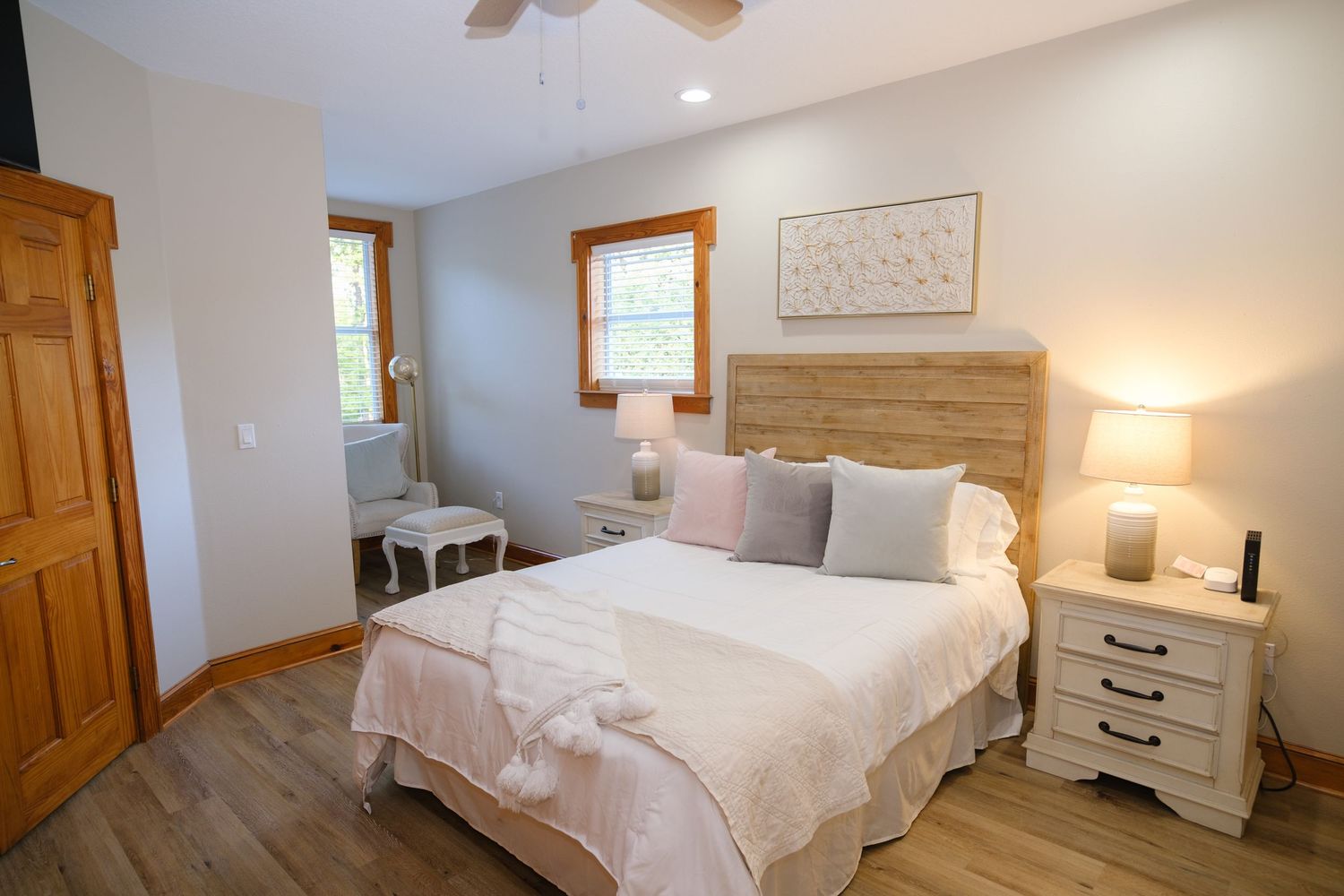 A cozy bedroom with rustic wood flooring, white bedding, and natural light streaming through wooden-framed windows.