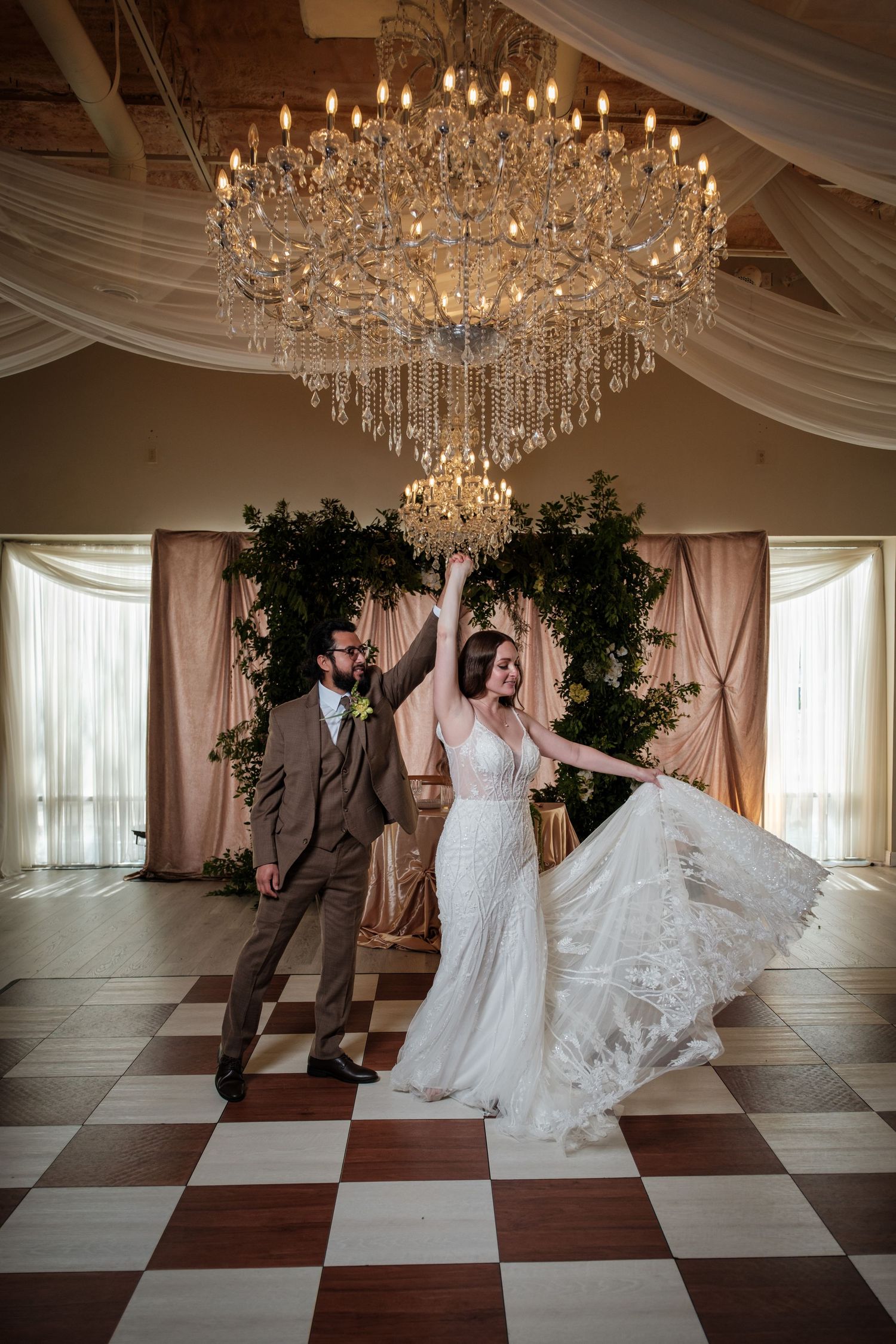 Bride and groom dancing under a crystal chandelier on a checkered floor at an elegant wedding reception venue.