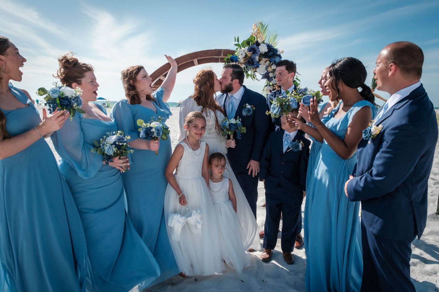 Beach wedding party celebrates newlyweds kissing under floral arch, bridesmaids in blue dresses with bouquets, flower girls nearby.
