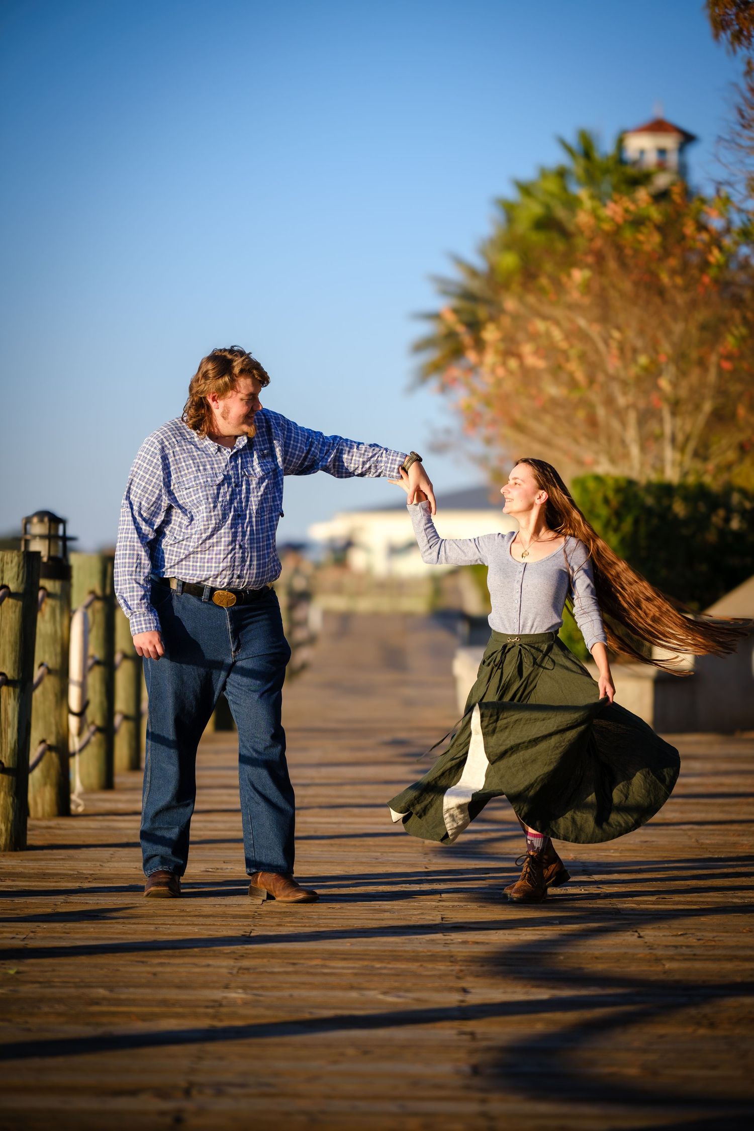 A couple dances joyfully on a wooden boardwalk at golden hour, with palm trees and blue sky in the background.