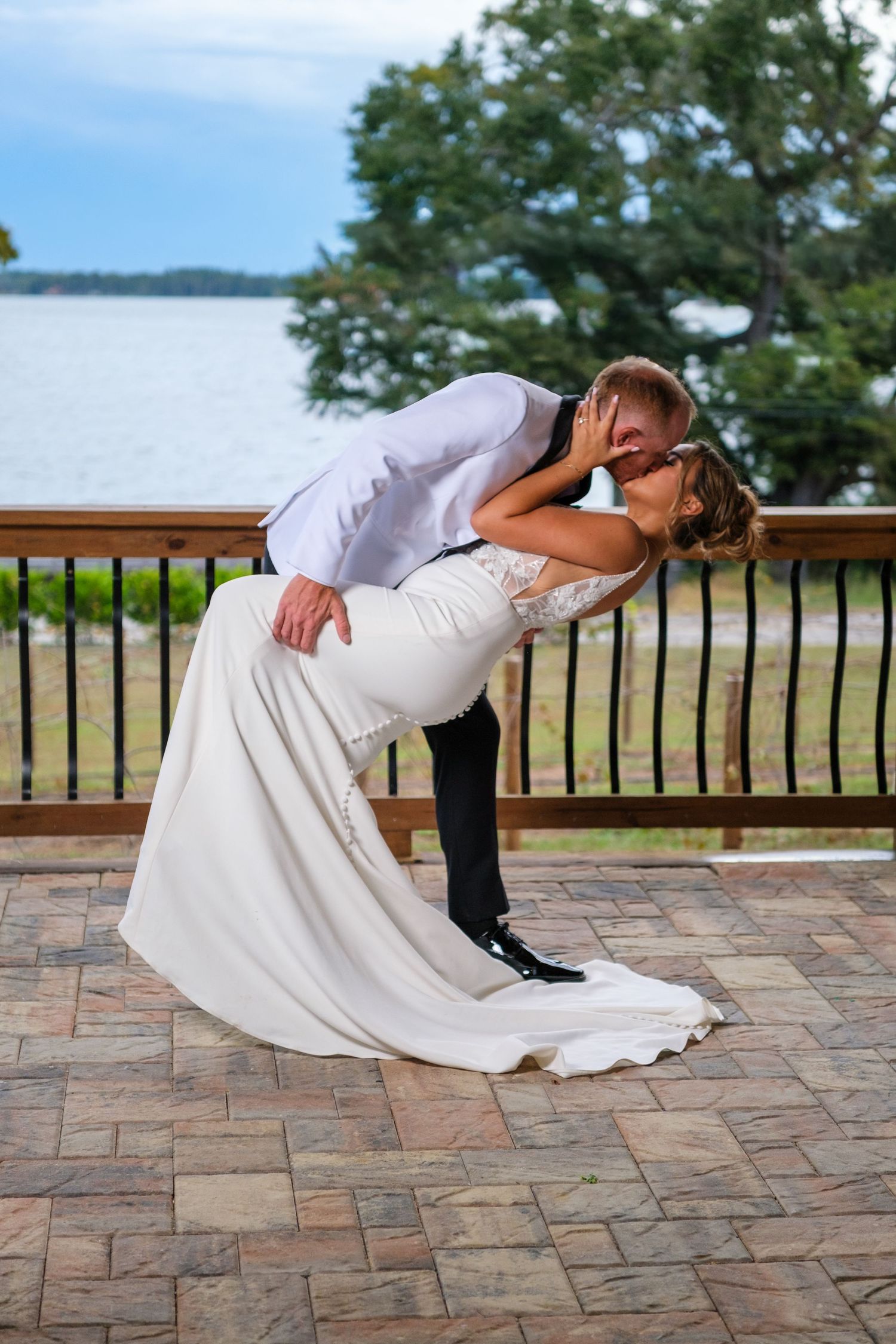 A romantic wedding couple performs a dramatic dip pose on a stone patio overlooking a waterfront view.