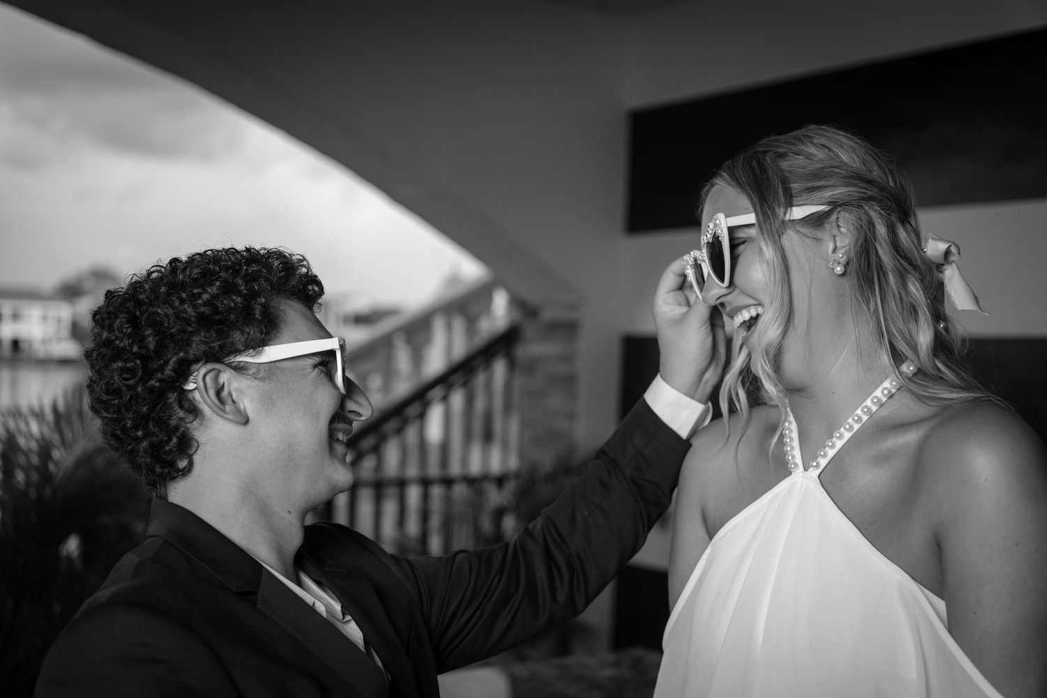 Black and white photo of a groom placing sunglasses on a laughing bride at their outdoor wedding.