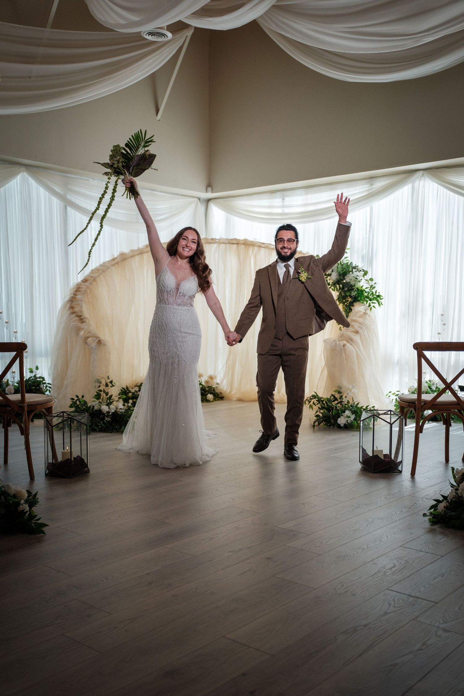 Joyful newlyweds celebrating after ceremony, bride in lace gown holding bouquet, groom in brown suit, elegant draped venue.