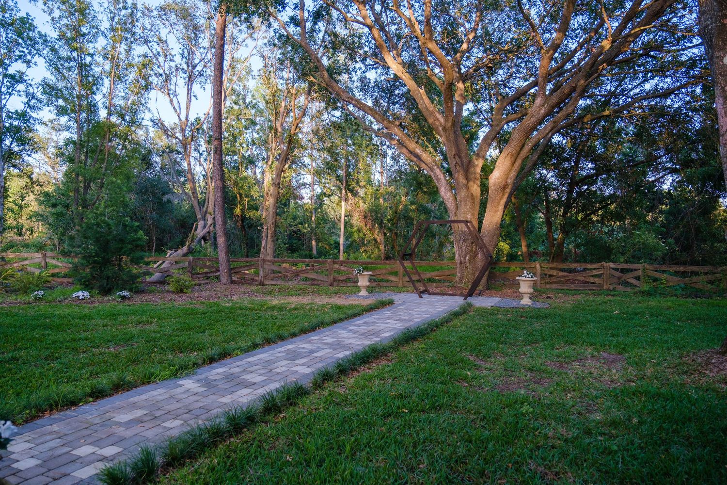 Stone pathway winding past garden urns and large trees in a peaceful woodland setting at dusk.