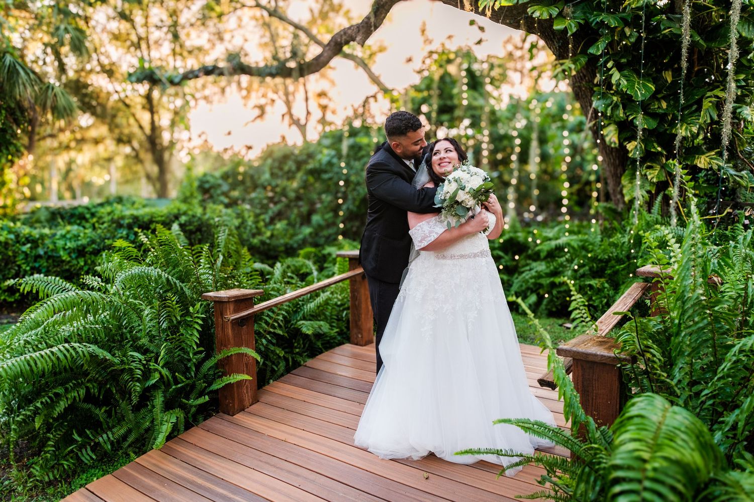 A couple shares a romantic moment on a wooden boardwalk surrounded by lush green ferns in a garden setting.