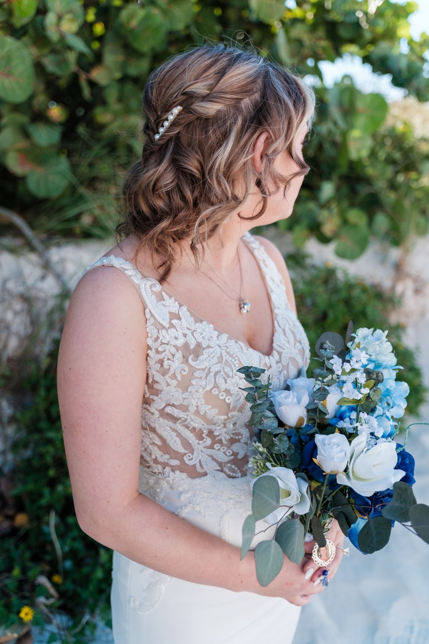 A wedding bouquet featuring light blue and white flowers with eucalyptus leaves against a lace dress.
