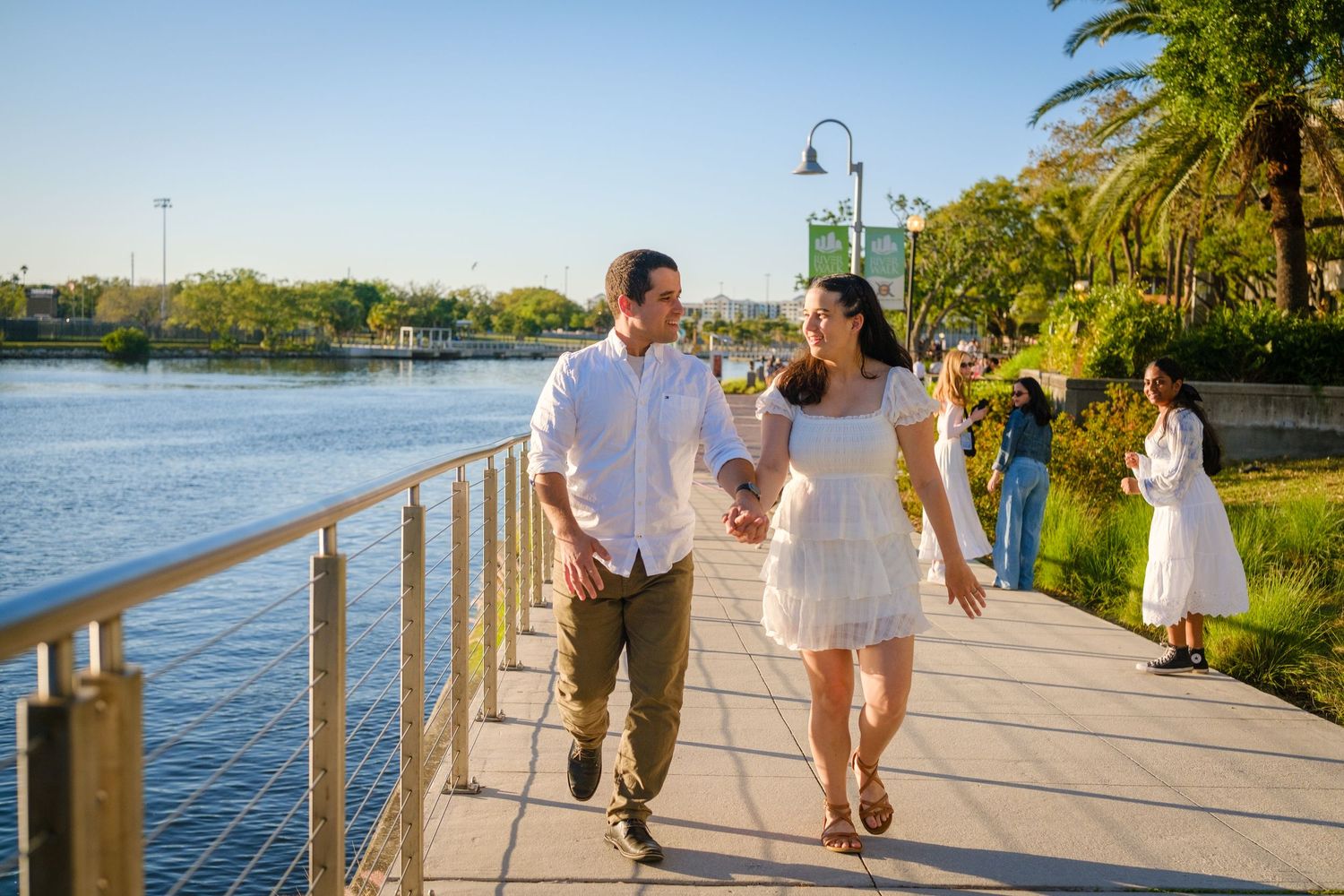 Couple holding hands walking along a scenic waterfront boardwalk surrounded by palm trees on a sunny day.