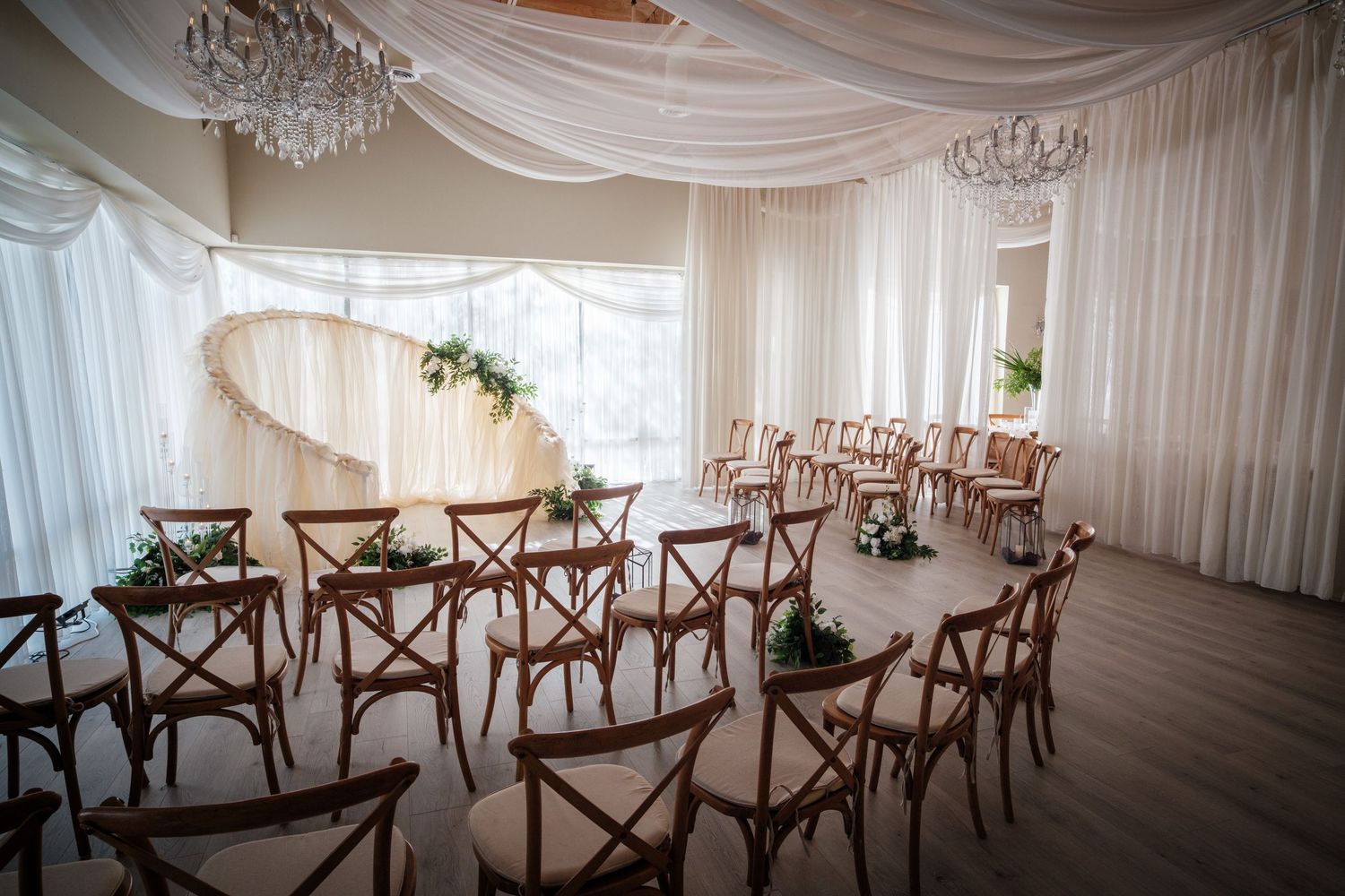 Elegant wedding ceremony venue with white draping, chandeliers, wooden cross-back chairs, and floral arrangements.