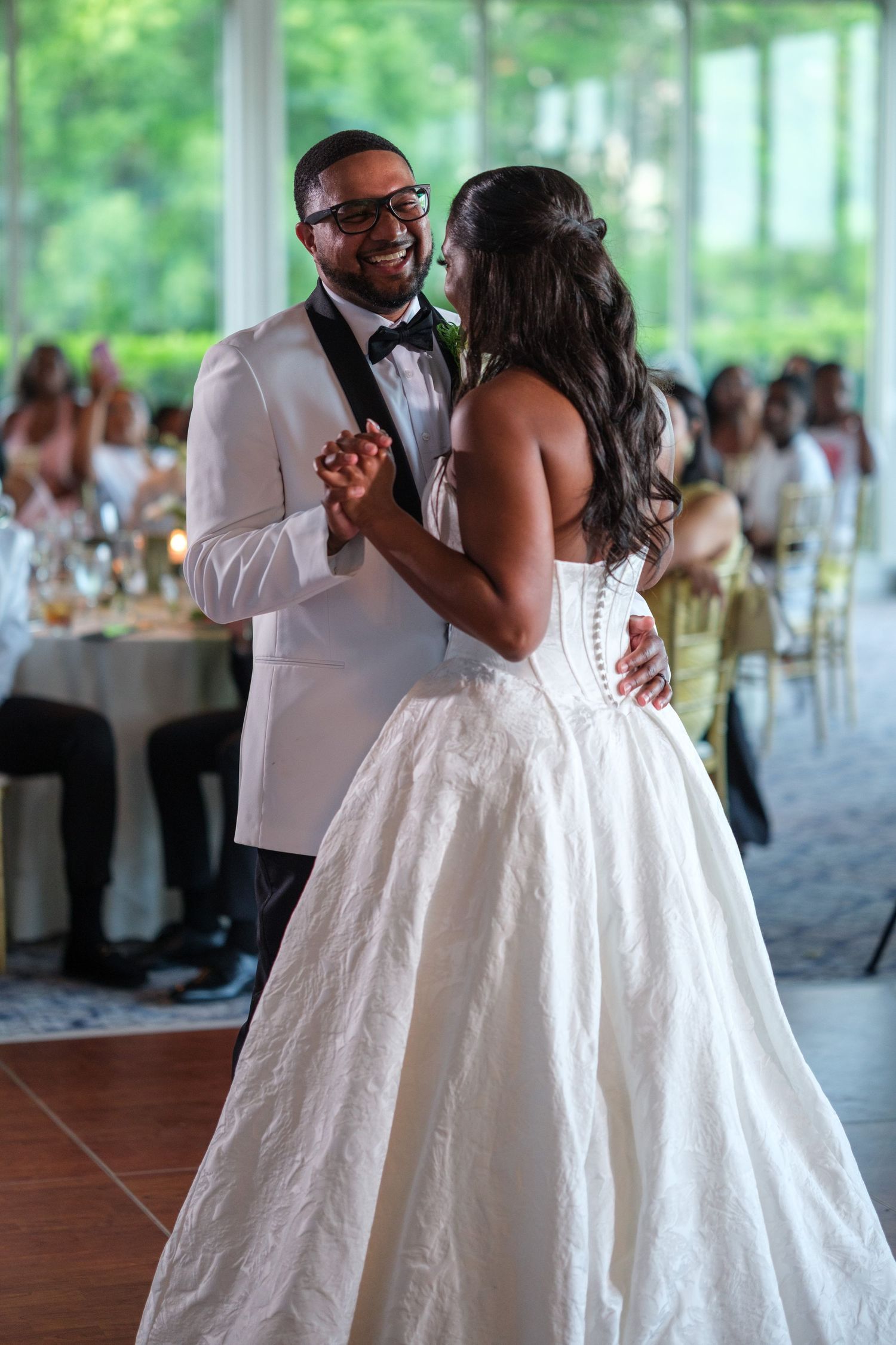 Bride and groom share their first dance, smiling joyfully, guests seated in background at wedding reception.