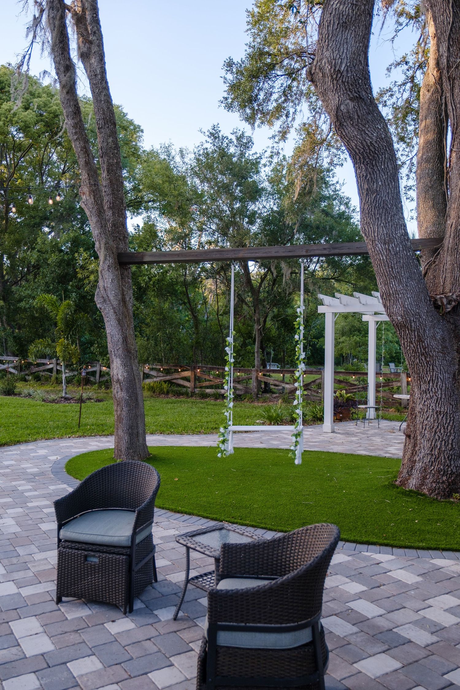 A tranquil outdoor seating area with wicker chairs and a swing set surrounded by mature trees on a paved patio.