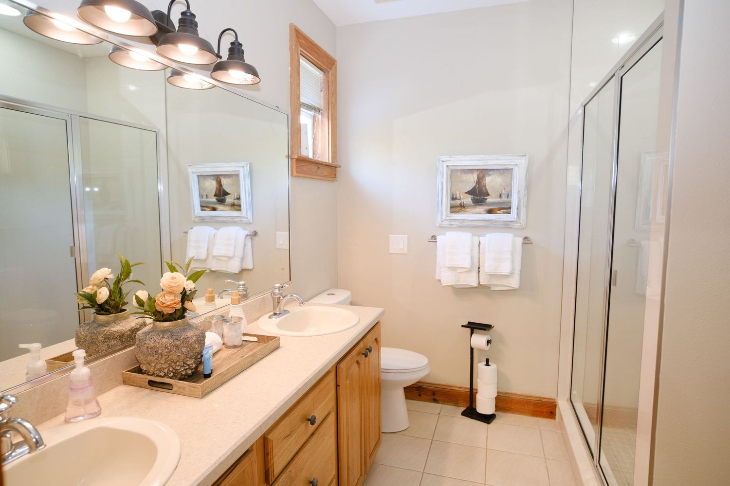 Modern bathroom interior with light wood vanity, dual sinks, large mirror, and decorative wall art.