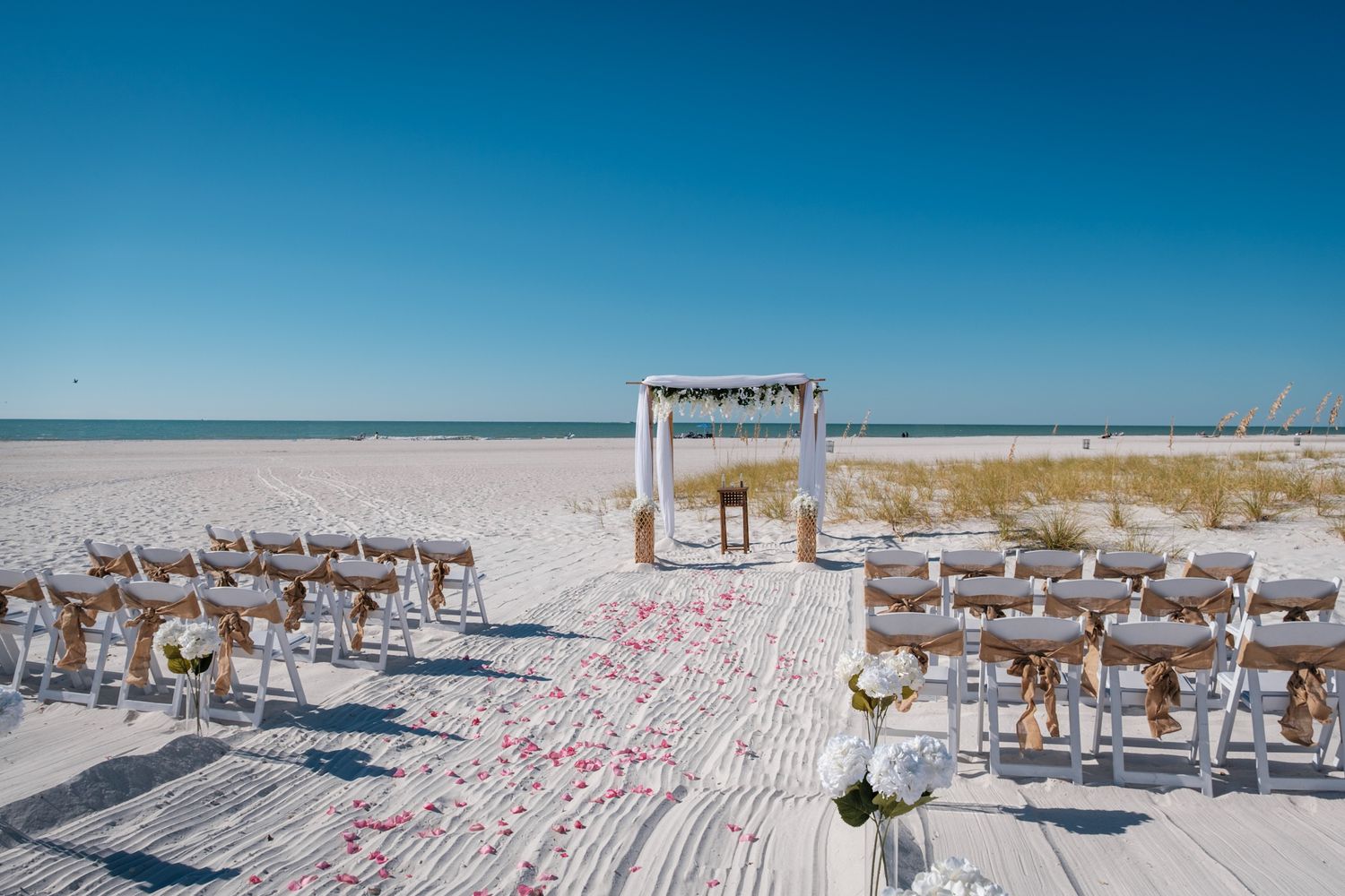 White wedding arch and chairs set up on pristine beach with pink rose petals scattered on white sand.
