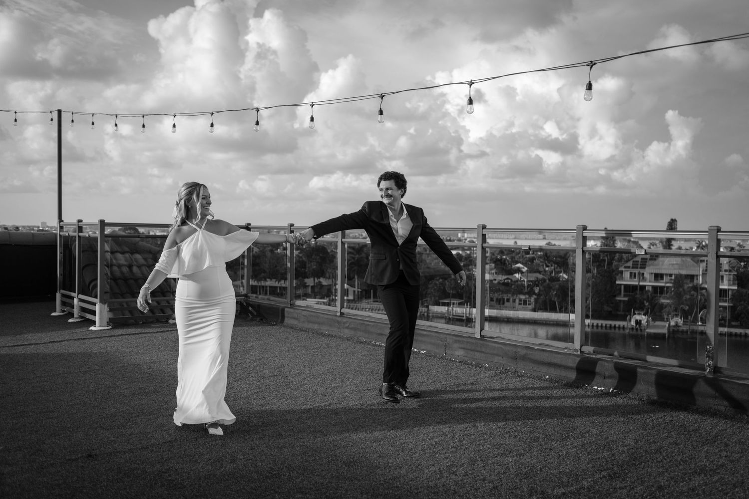 Black and white photo of a bride and groom dancing on a rooftop terrace with string lights and cloudy sky.