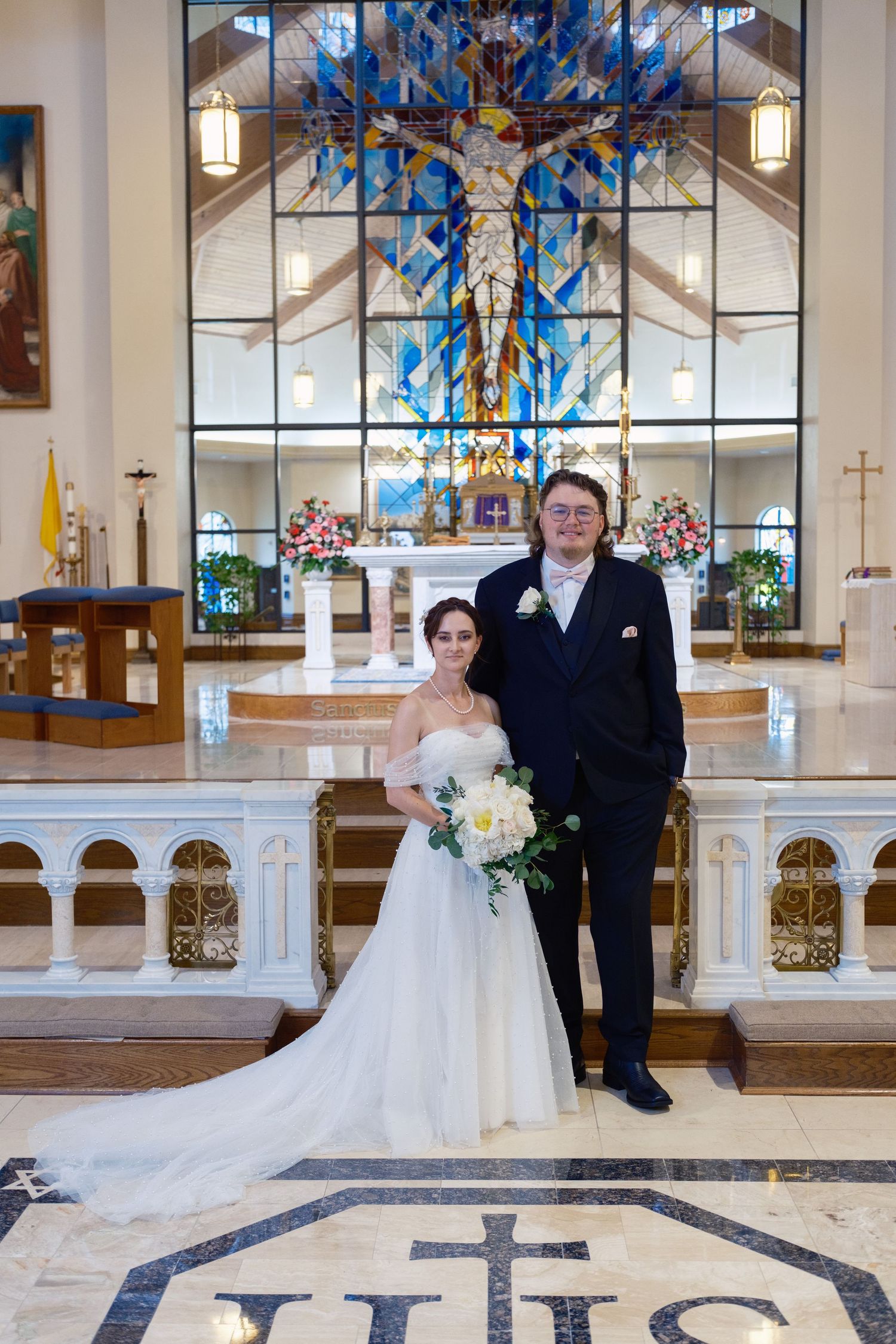 Bride and groom posing at church altar with stunning blue stained glass window backdrop on their wedding day.