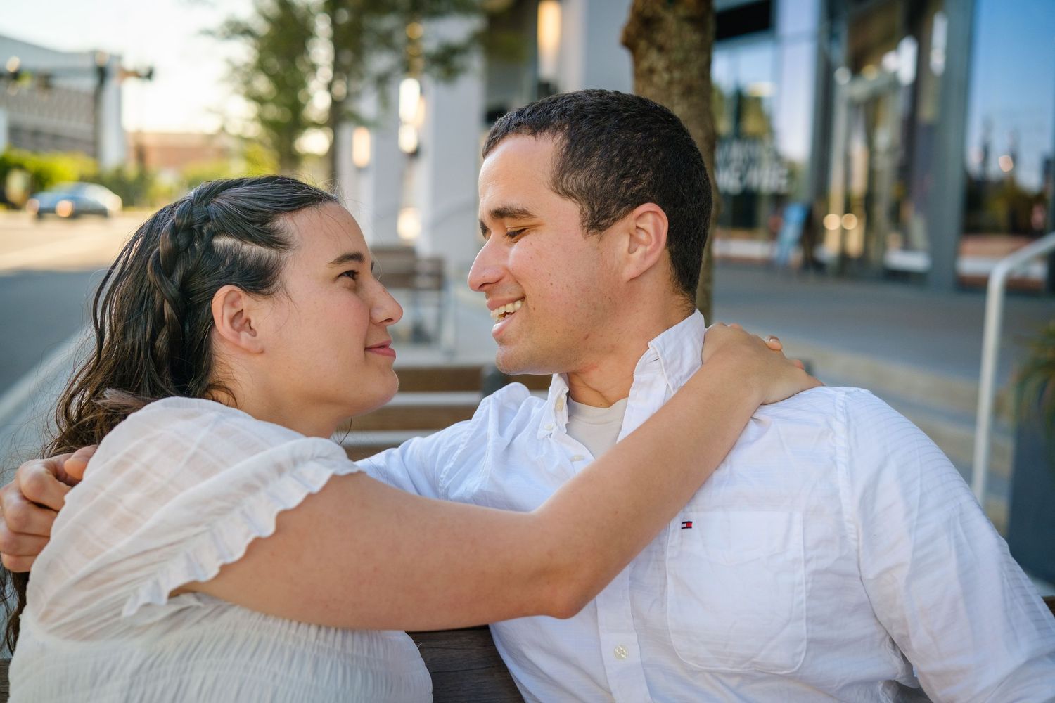 A couple in white outfits share a romantic moment outdoors, smiling at each other while embracing on a bench.