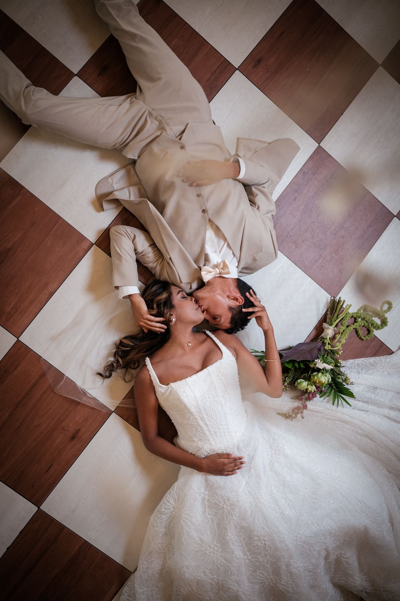 Bride and groom lying on checkered floor kissing, aerial view of wedding couple in white dress and beige suit.