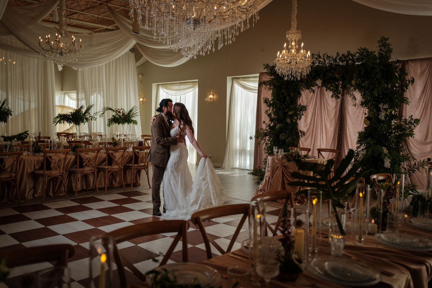 Bride and groom share romantic dance in elegant ballroom with chandelier, draped ceilings, and lush greenery arch.