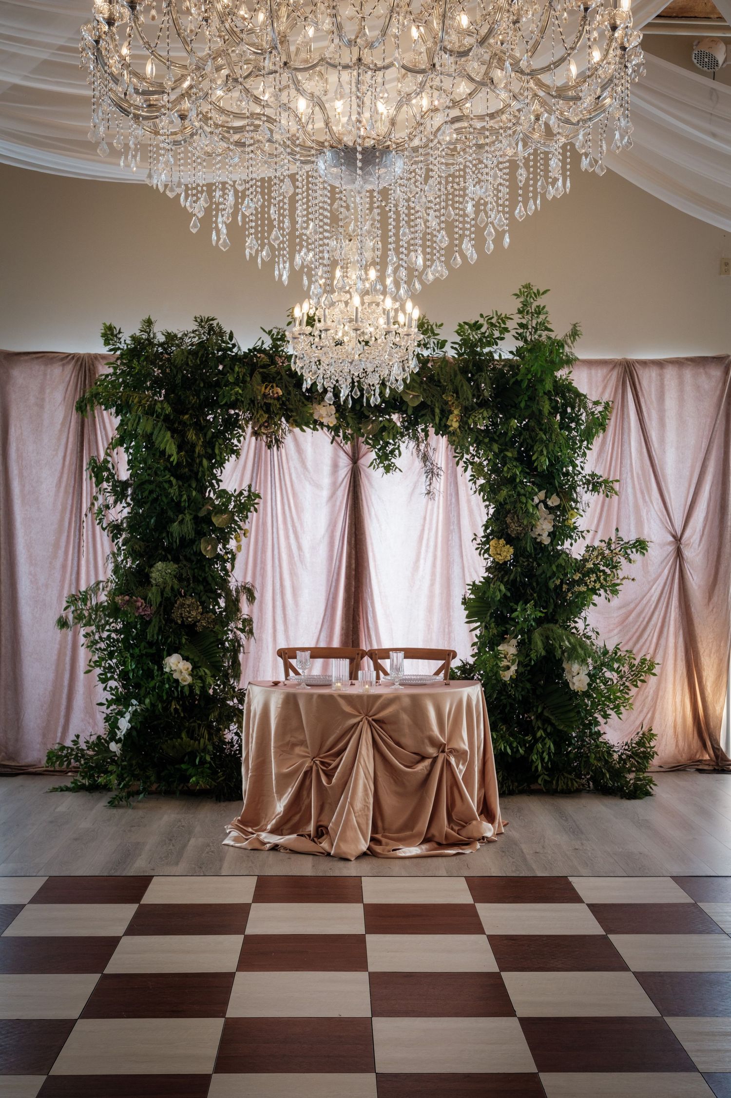 Elegant wedding sweetheart table with gold satin cloth, greenery arch, pink drapes, and crystal chandelier overhead.