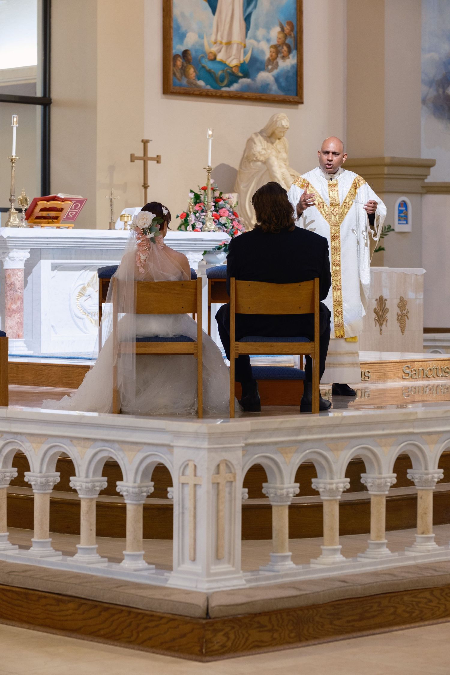 Bride and groom seated before priest during Catholic wedding ceremony at church altar.
