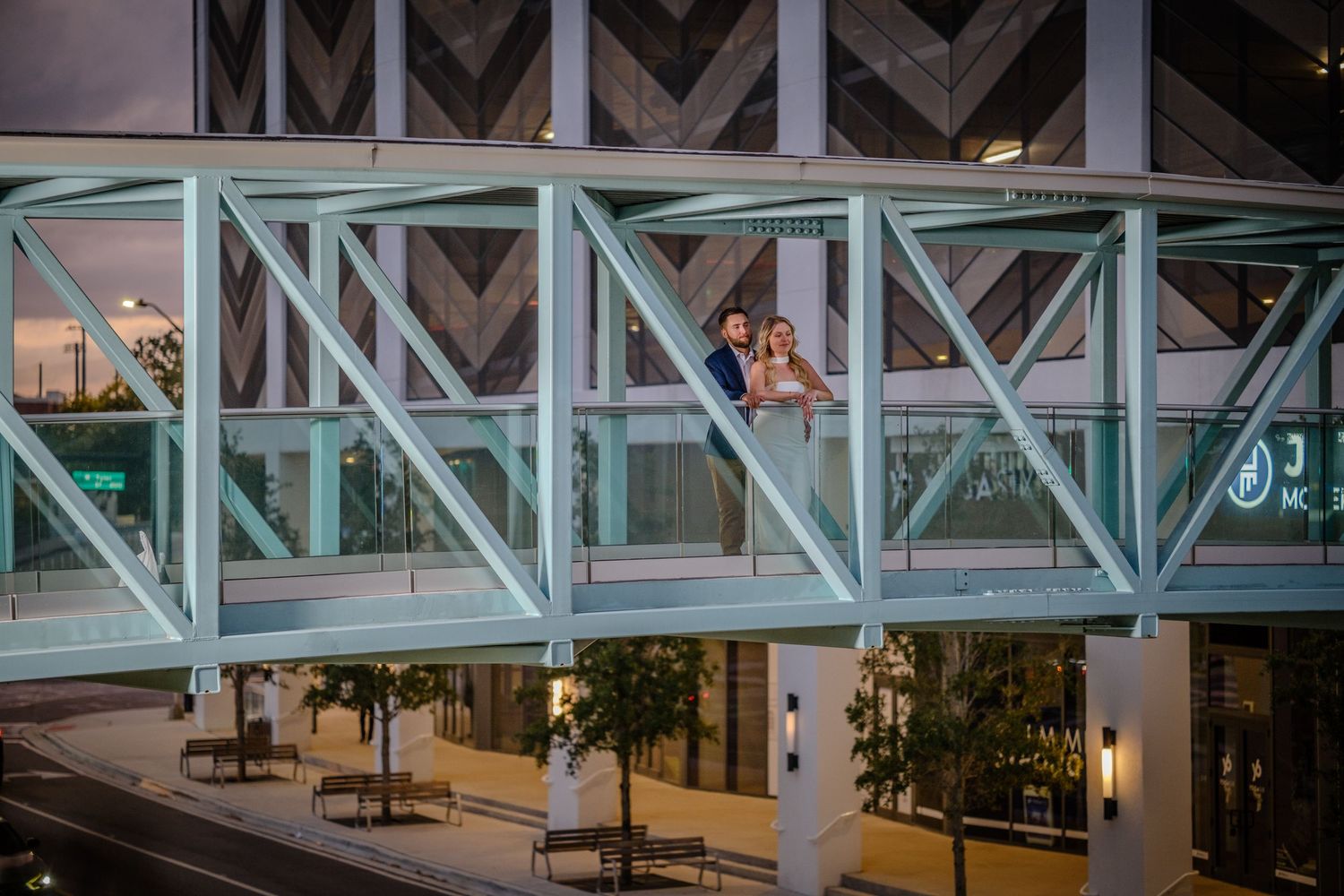 Couple posing on a modern glass and steel skybridge walkway at dusk, surrounded by city architecture.