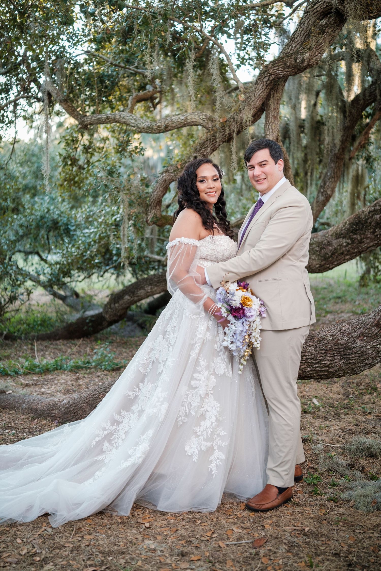 A couple in wedding attire poses together under a large oak tree draped with Spanish moss in a romantic outdoor setting.
