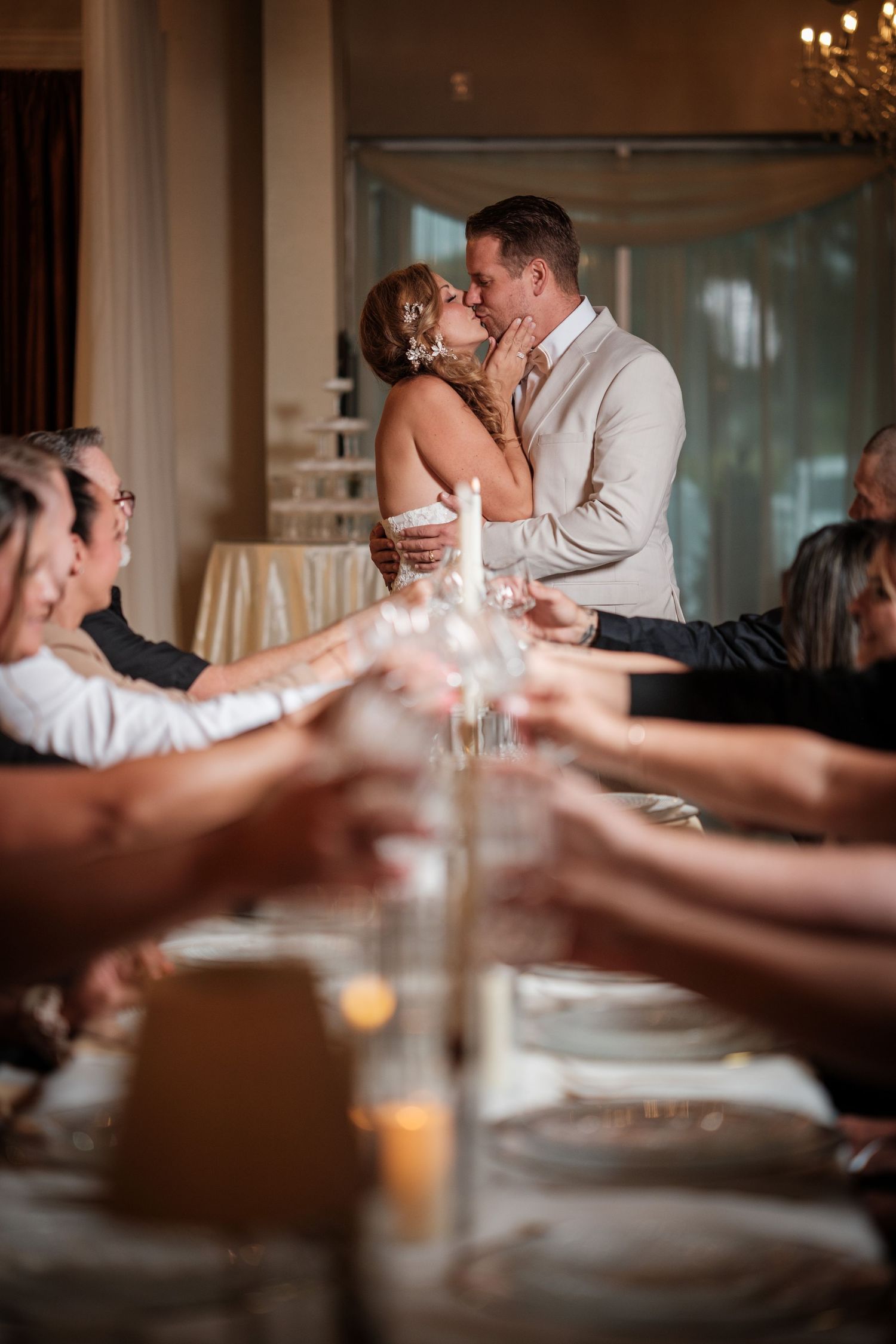 Bride and groom share a romantic kiss at wedding reception dinner table surrounded by guests raising glasses in a toast.
