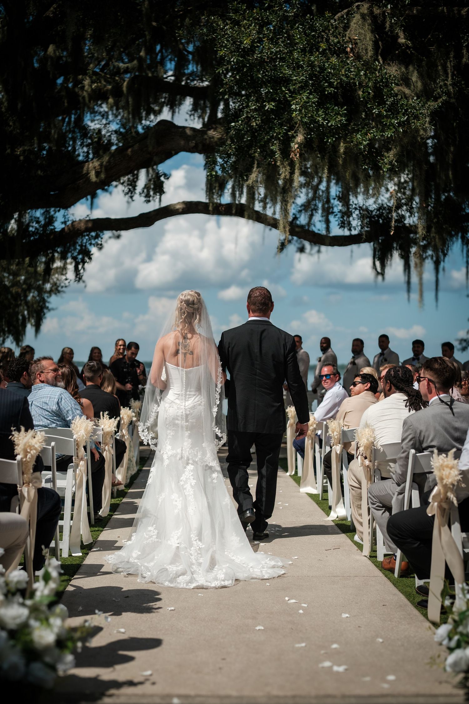 Bride and groom walk down outdoor wedding aisle under moss-draped oak tree with guests seated on white chairs.