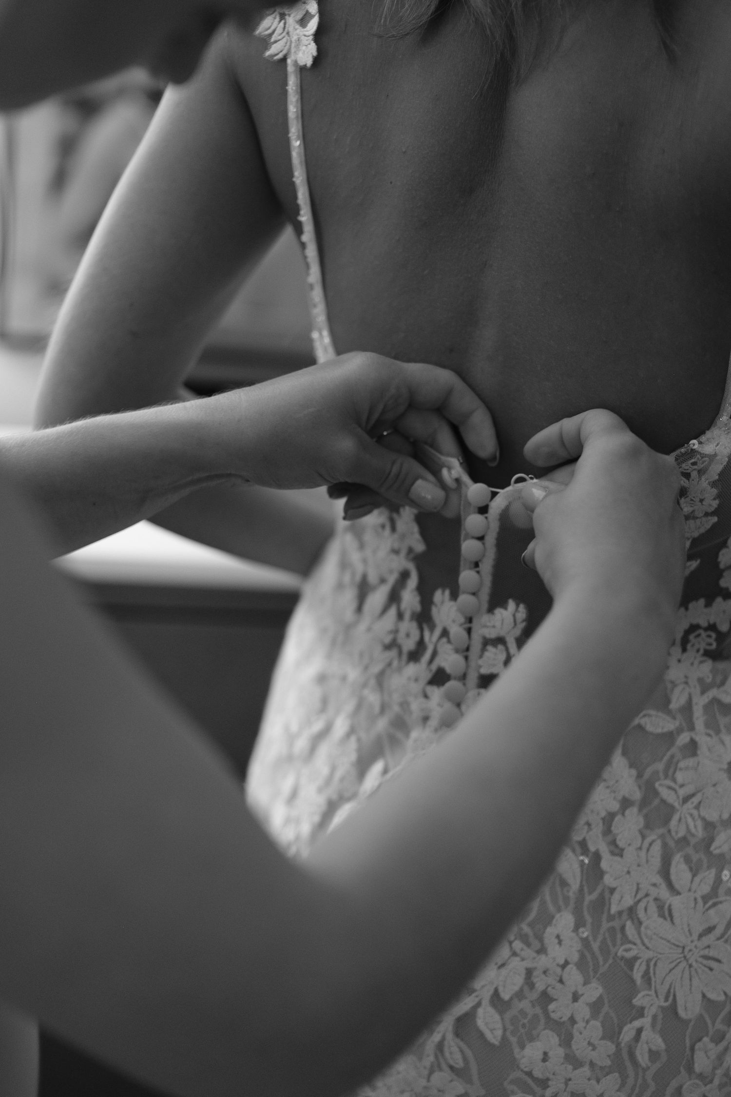 A close-up black and white photo showing hands adjusting the lacing on the back of a delicate lace wedding dress.