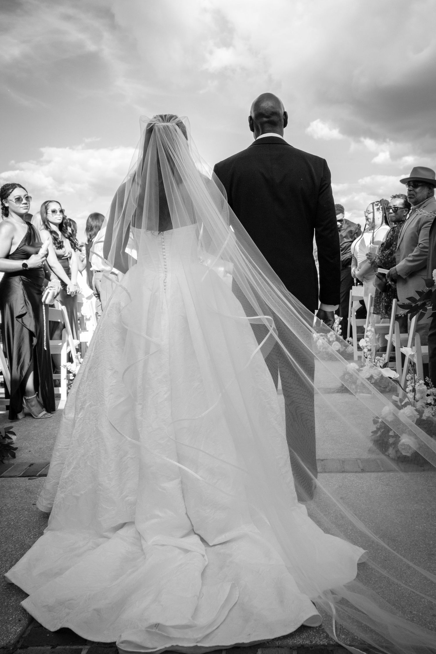 Black and white photo of bride in flowing veil and gown walking down aisle with groom at outdoor wedding.