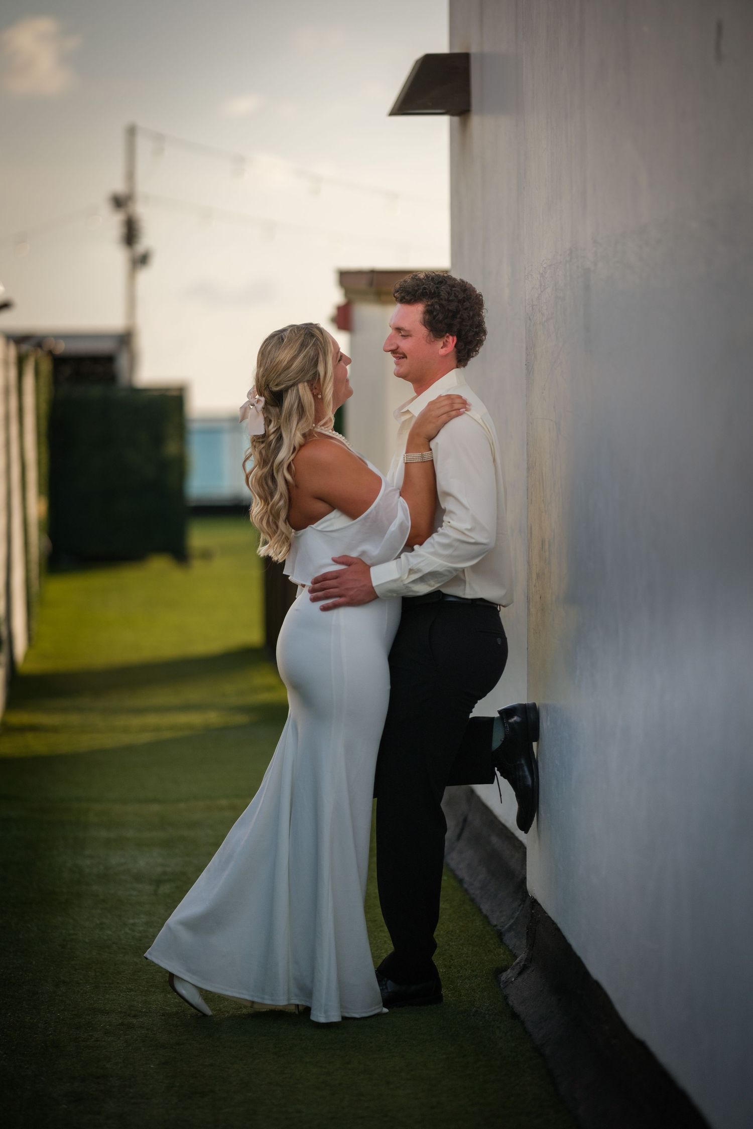 A couple in wedding attire embracing and smiling against a white wall outdoors at dusk.