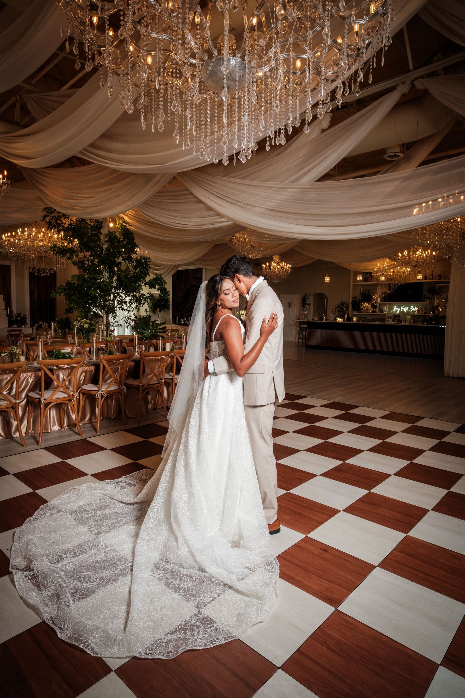 Bride and groom share an intimate embrace on a checkered dance floor beneath sparkling chandeliers in a decorated ballroom.