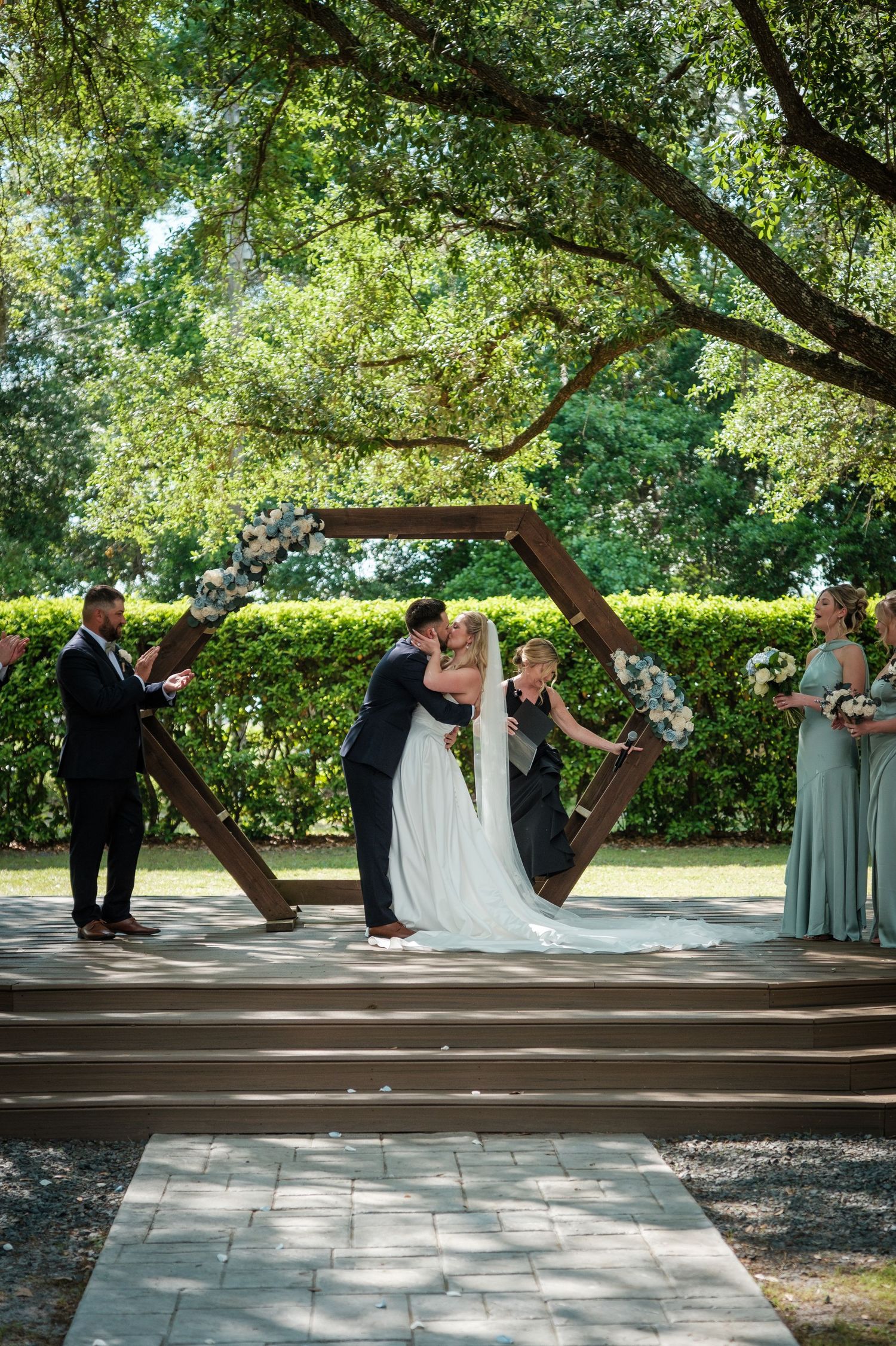 Couple shares first kiss under hexagonal wooden arch adorned with white flowers at an outdoor garden wedding ceremony.