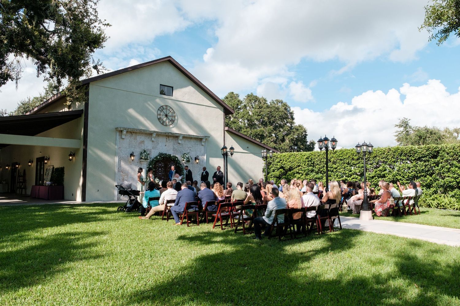 A panoramic view of a wedding ceremony setup outdoors with white chairs arranged on a sunny lawn near a building.