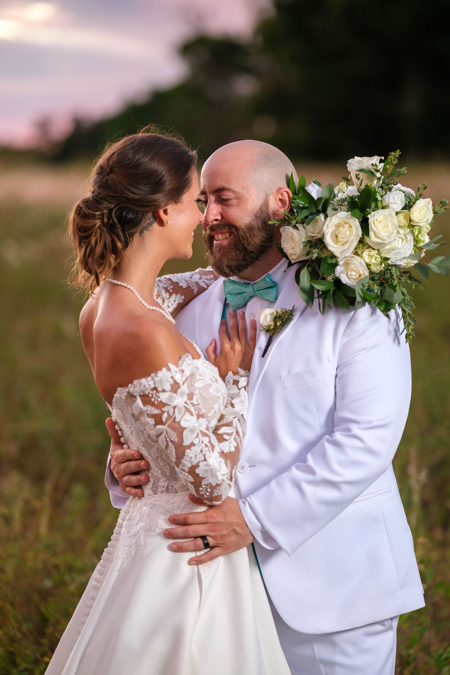A romantic wedding couple shares a tender embrace in a field at sunset with white floral bouquet details.