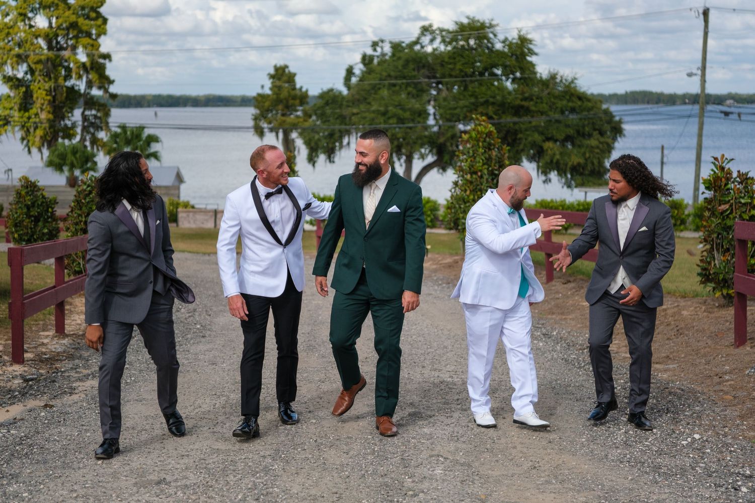 A group of people in formal attire laughing together on a waterfront path with trees and a lake in the background.