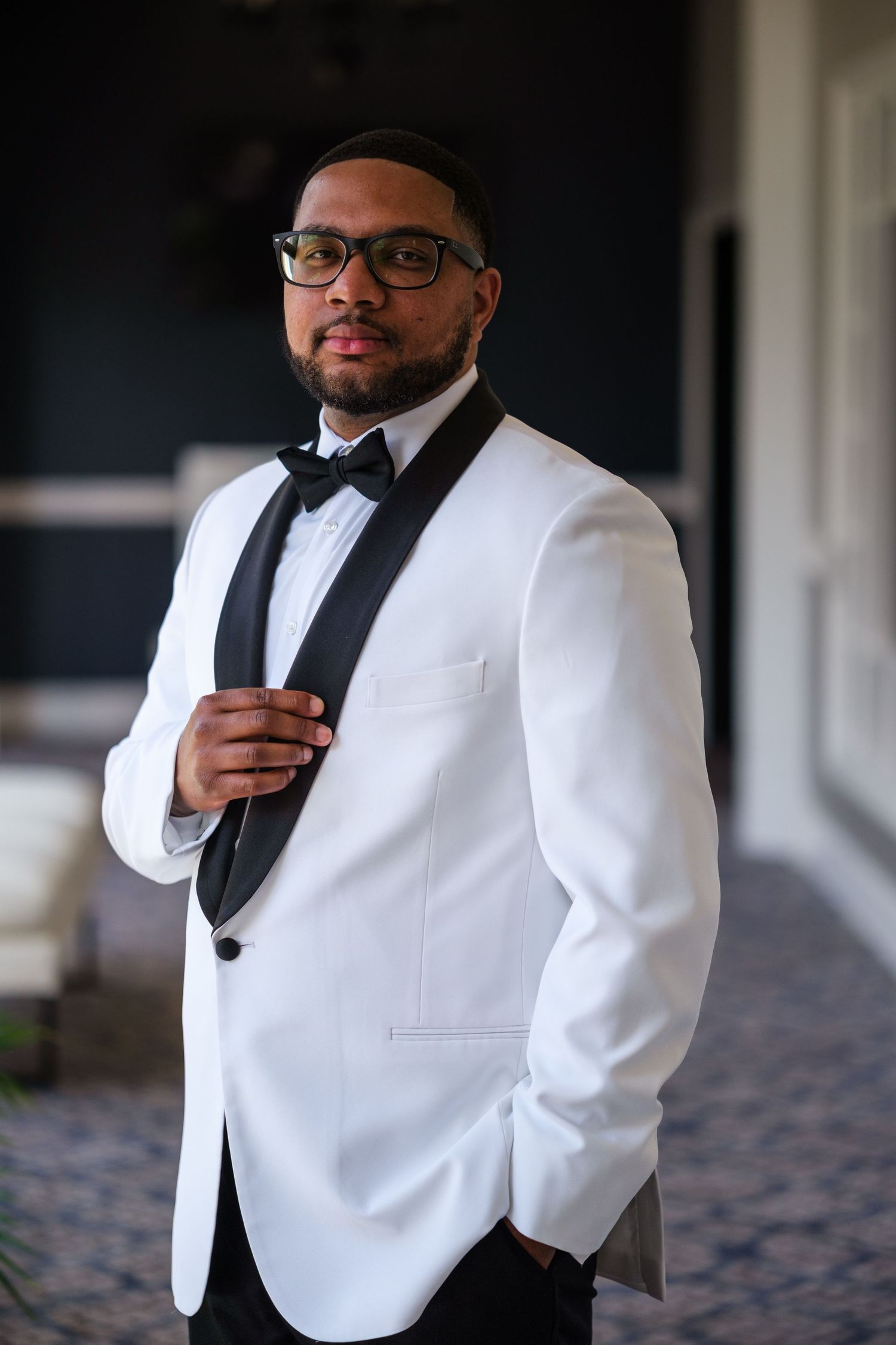 A well-dressed man in a white tuxedo jacket with black lapels and bow tie, wearing glasses, posing confidently.