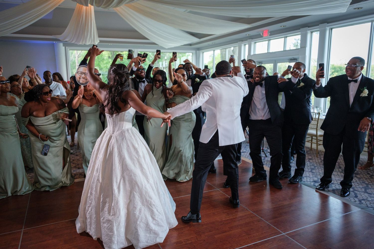 Bride and groom share first dance surrounded by cheering bridesmaids in sage green gowns and groomsmen at wedding reception.
