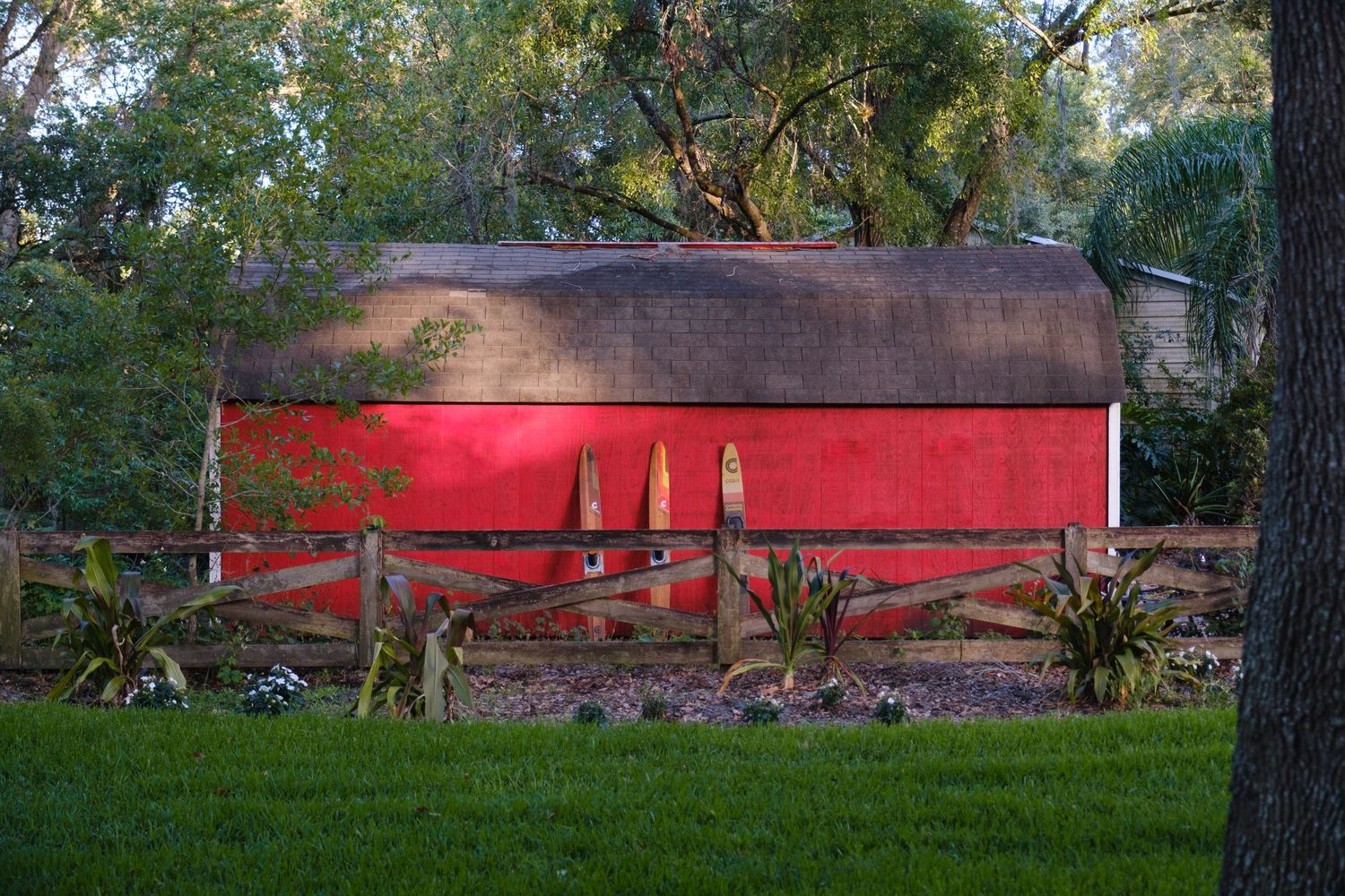 A bright red barn with a rustic brown roof stands among trees and green grass at dusk.