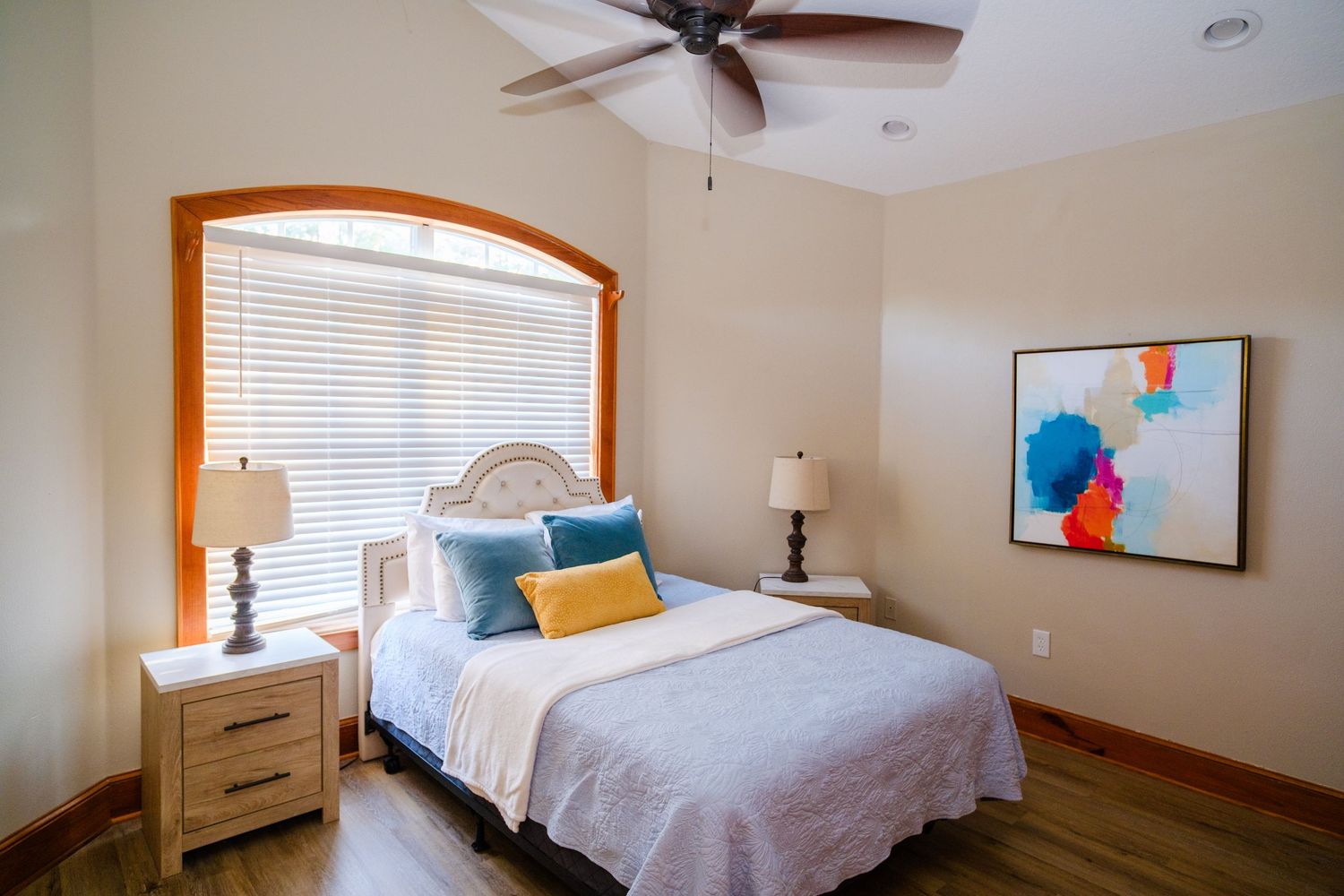 A cozy bedroom with white bedding, wooden window trim, and colorful abstract art on the wall features a ceiling fan.