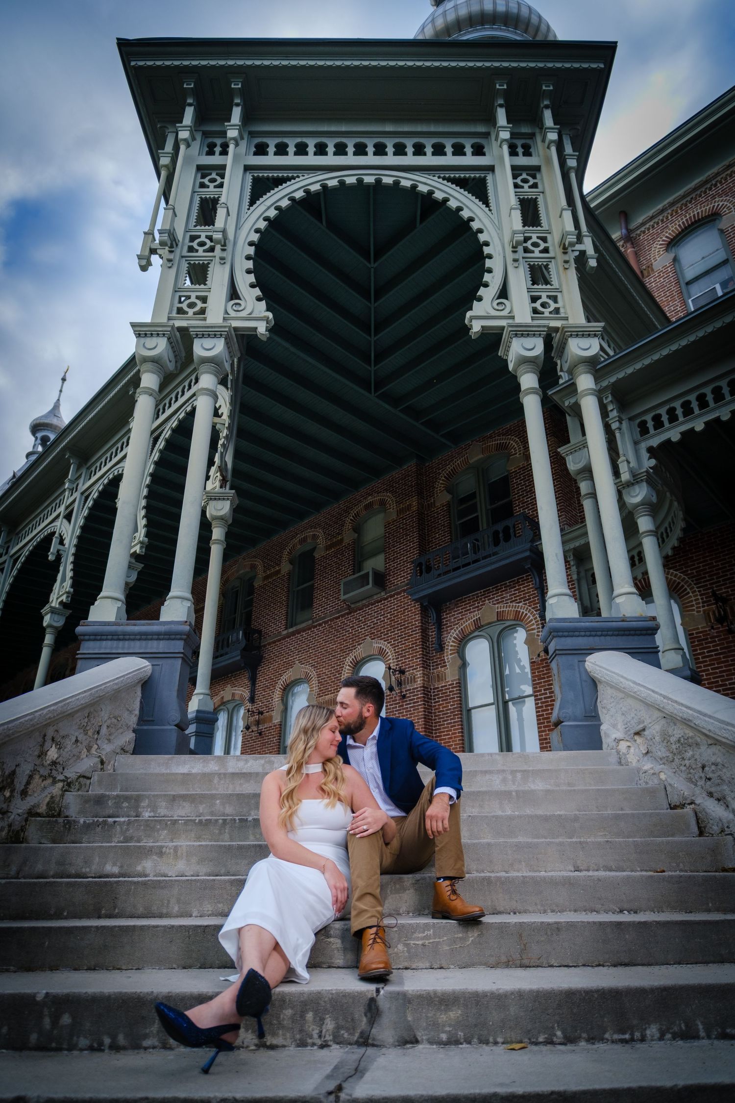 Couple sitting on ornate Victorian building steps, woman in white dress, man in blue blazer, romantic engagement photo.