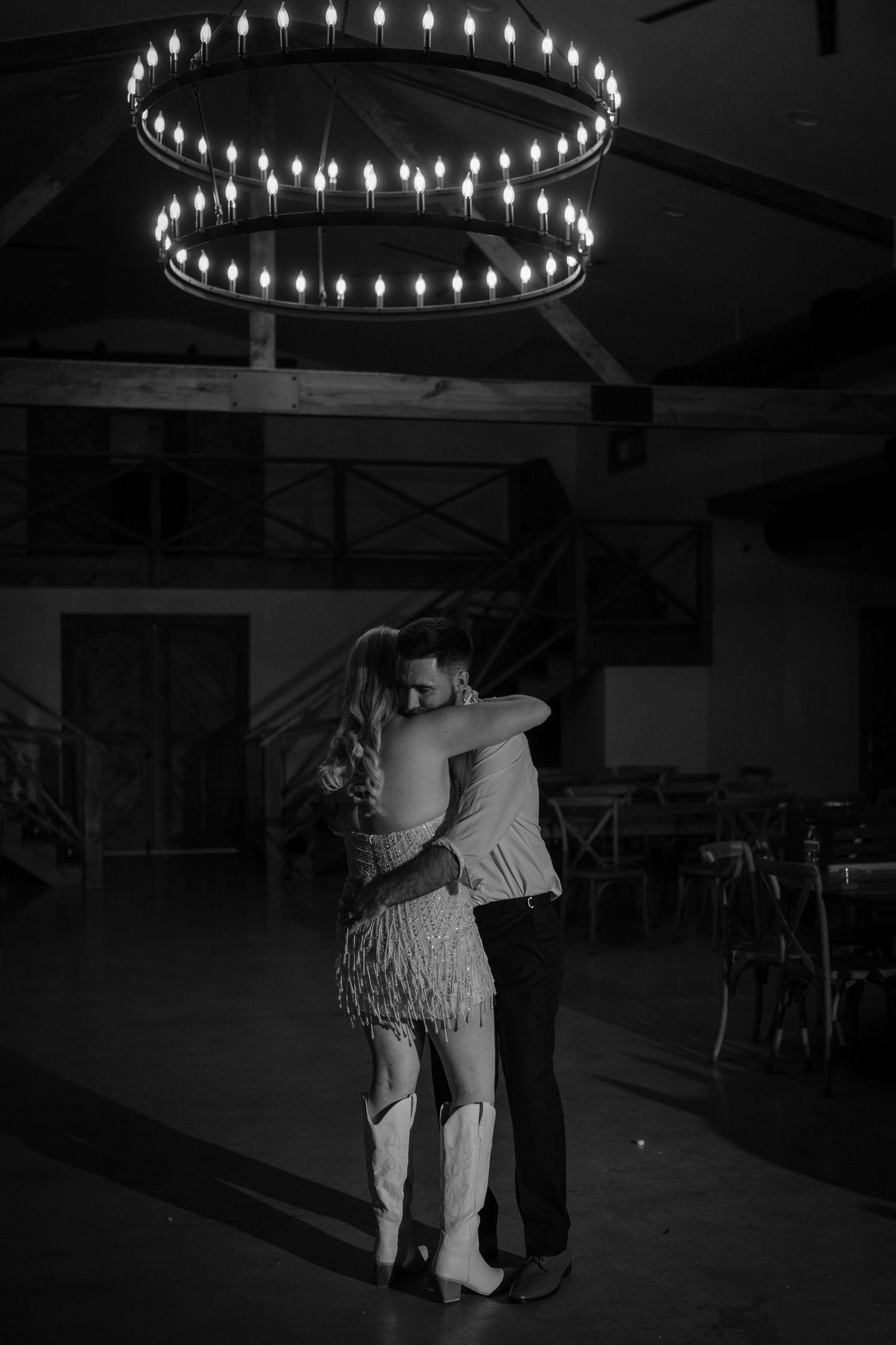 Black and white photo of a couple slow dancing under a circular chandelier in a dimly lit wedding venue.