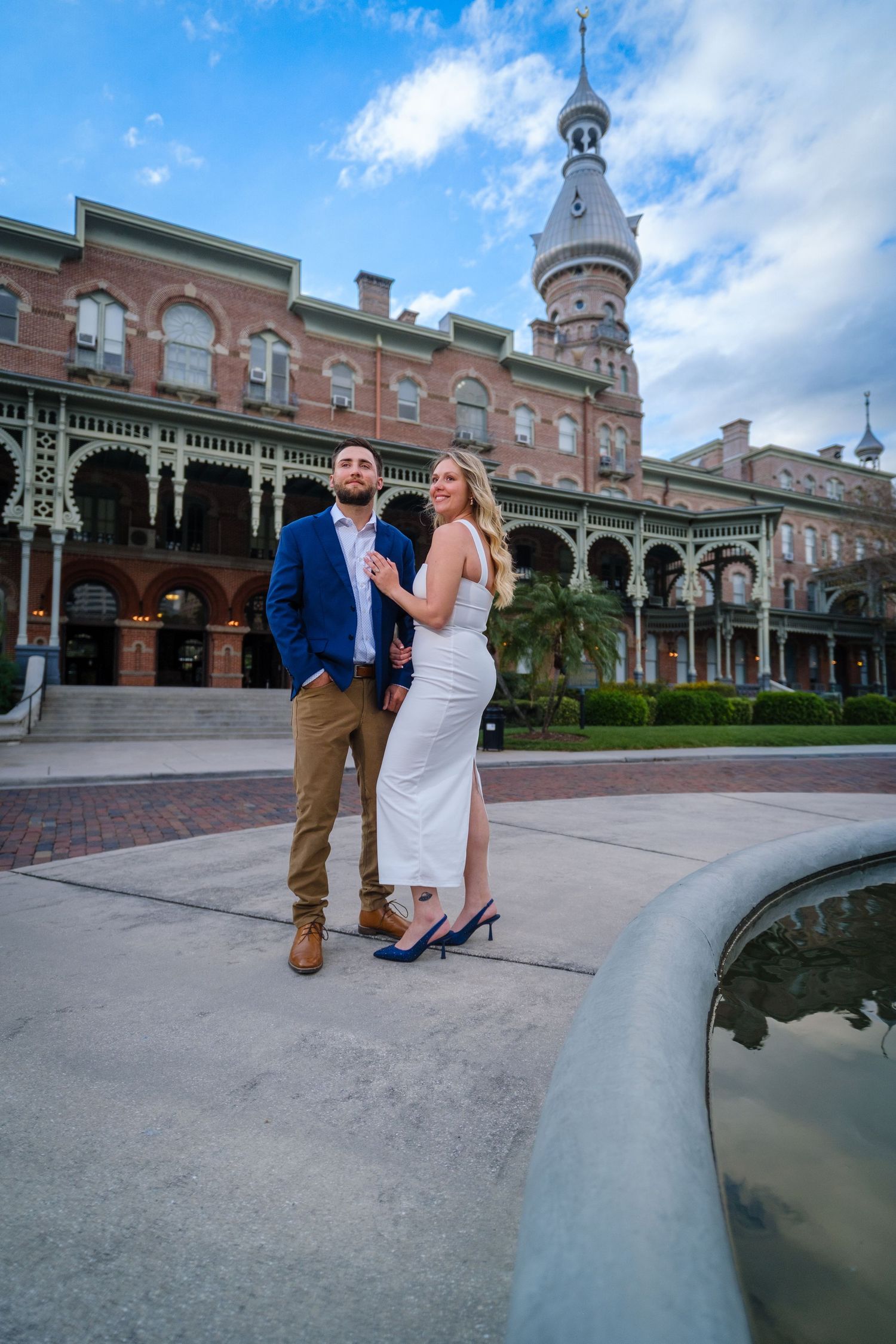 A couple poses for romantic photos in front of a historic Victorian-style building with ornate architecture and a reflecting pool.