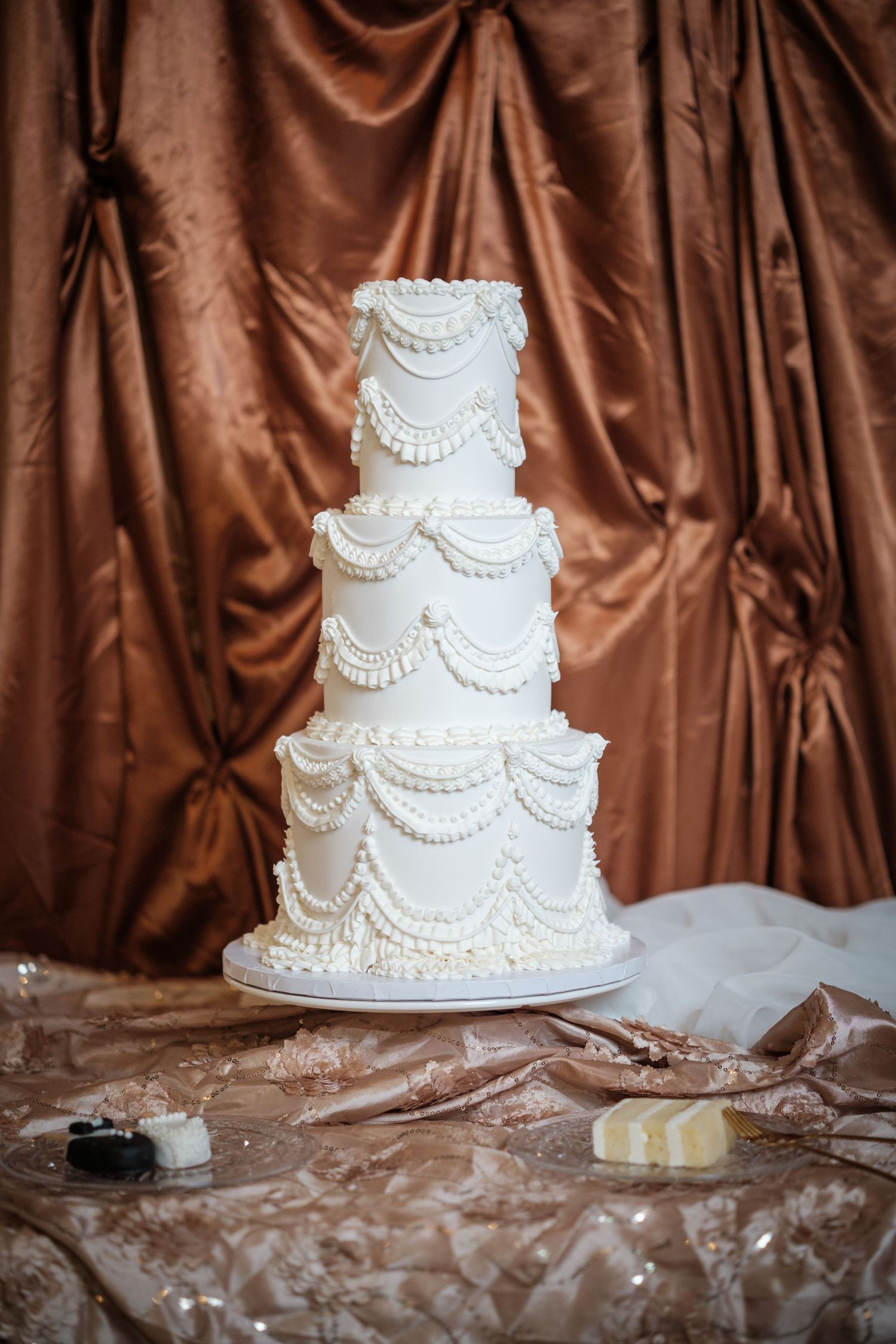 Elegant four-tier white wedding cake with pearl and swag piping details on a decorated table with brown satin backdrop.