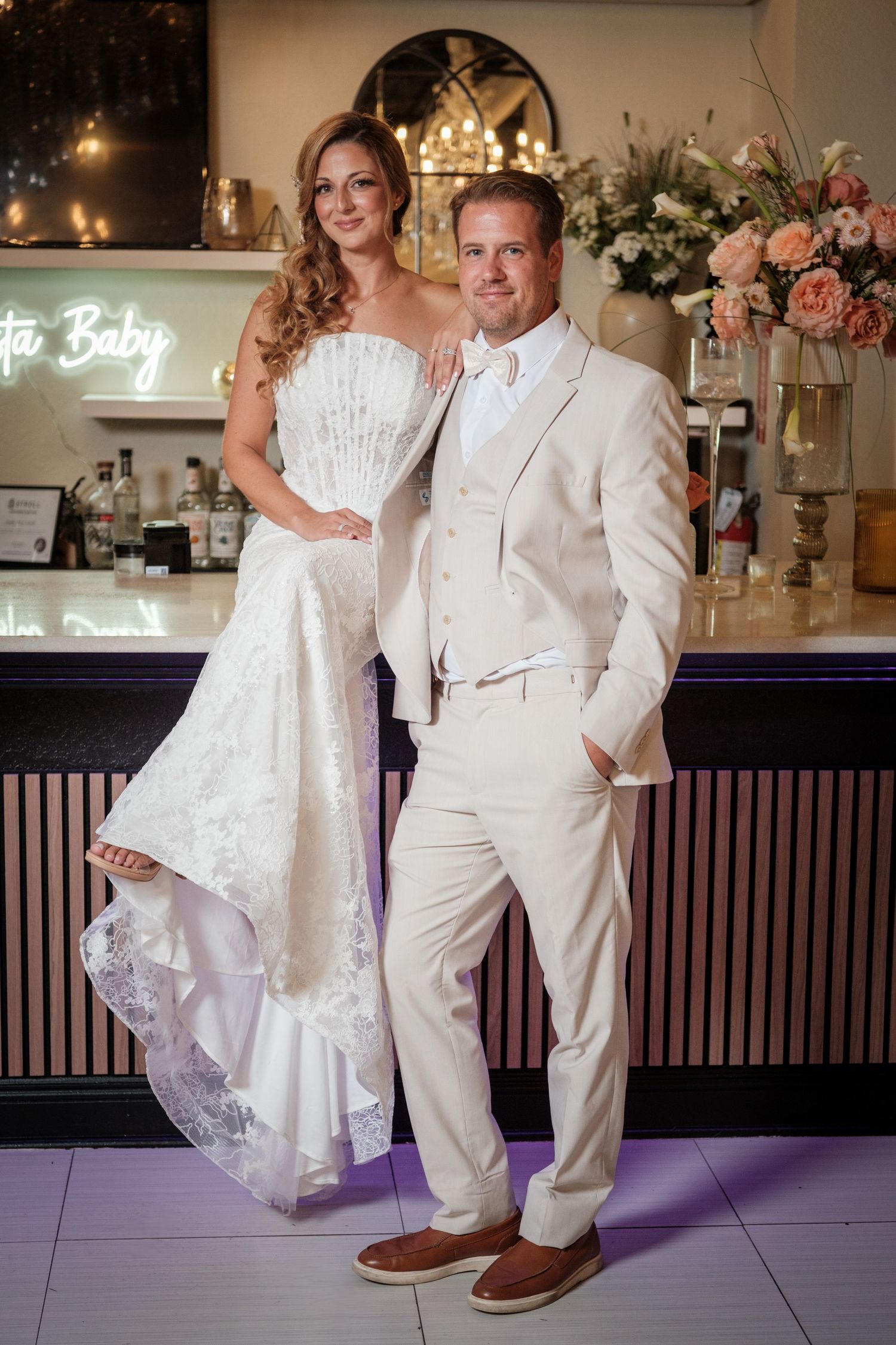 Elegant bride in strapless lace gown and groom in cream suit pose together at a stylish wedding venue bar.
